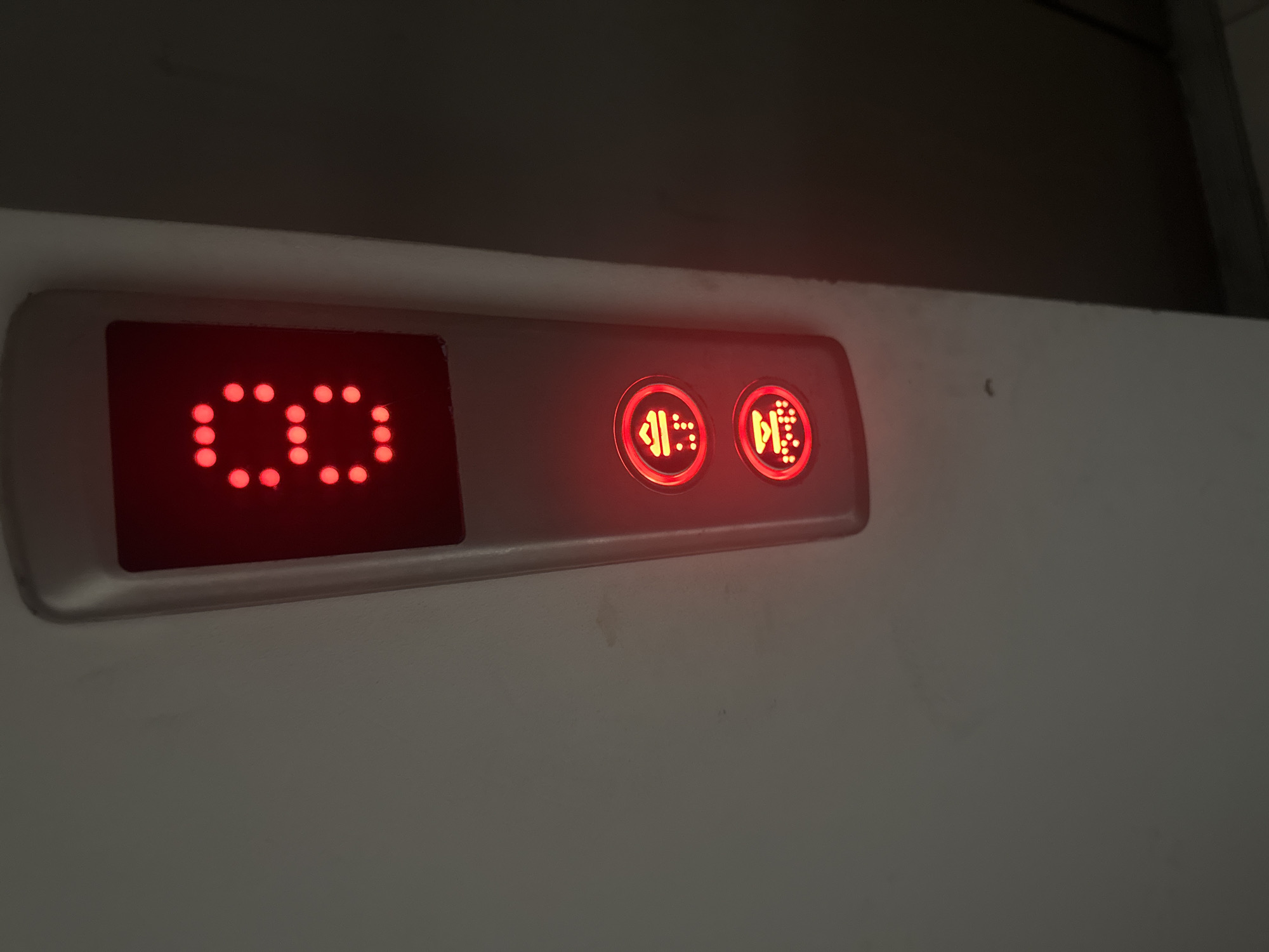 The image shows an elevator panel with a red digital display showing the number "8". Below the number display are two red illuminated buttons, each with a pictogram and braille. The top button has a pictogram of an elevator car with an upward arrow and braille. The bottom button has a pictogram of an elevator car with a downward arrow and braille. The elevator panel is mounted on a light-colored wall. The background is dark, suggesting the photo was taken inside an elevator or a dimly lit hallway. There is no visible indication of the location in Tbilisi, Georgia.