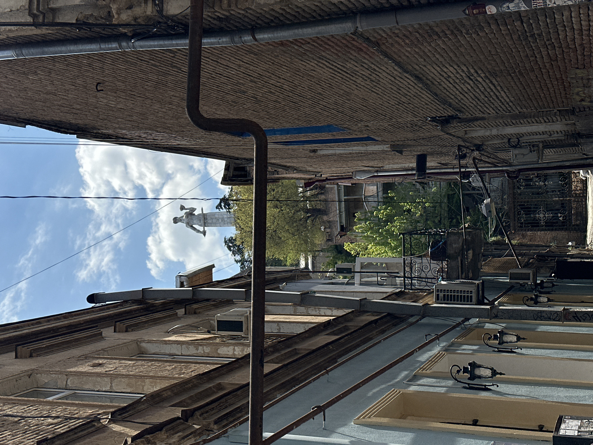 The image depicts a narrow urban passage, likely an alley or street, lined by old, multi-story buildings, under a bright, partly cloudy sky. The perspective looks towards the sky and a distant monument. On the left, an aged building in earthy tones features multiple windows with decorative cornices and an air conditioning unit near the roof. On the right, another building with a light blue section displays two ornate, dark metal wall sconces, windows, and intricate wrought-iron elements on balconies and railings further back.
A prominent, thick dark metal pipe runs vertically along the right building's facade and then bends horizontally across the upper frame. Numerous thinner pipes and electrical wires crisscross the space between the structures. In the distance, silhouetted against the bright blue sky, a large, light-colored statue of a male figure, possibly holding a horn or an object aloft, stands on a tall pedestal or rooftop, flanked by green trees. The brick pattern of an overhang is visible directly above the viewer's perspective. The scene is illuminated by clear daylight, suggesting midday. Faint, unreadable text or stickers are visible high on a distant wall. The setting is characteristic of an old quarter in Tbilisi, Georgia, with its dense architecture and exposed infrastructure.
