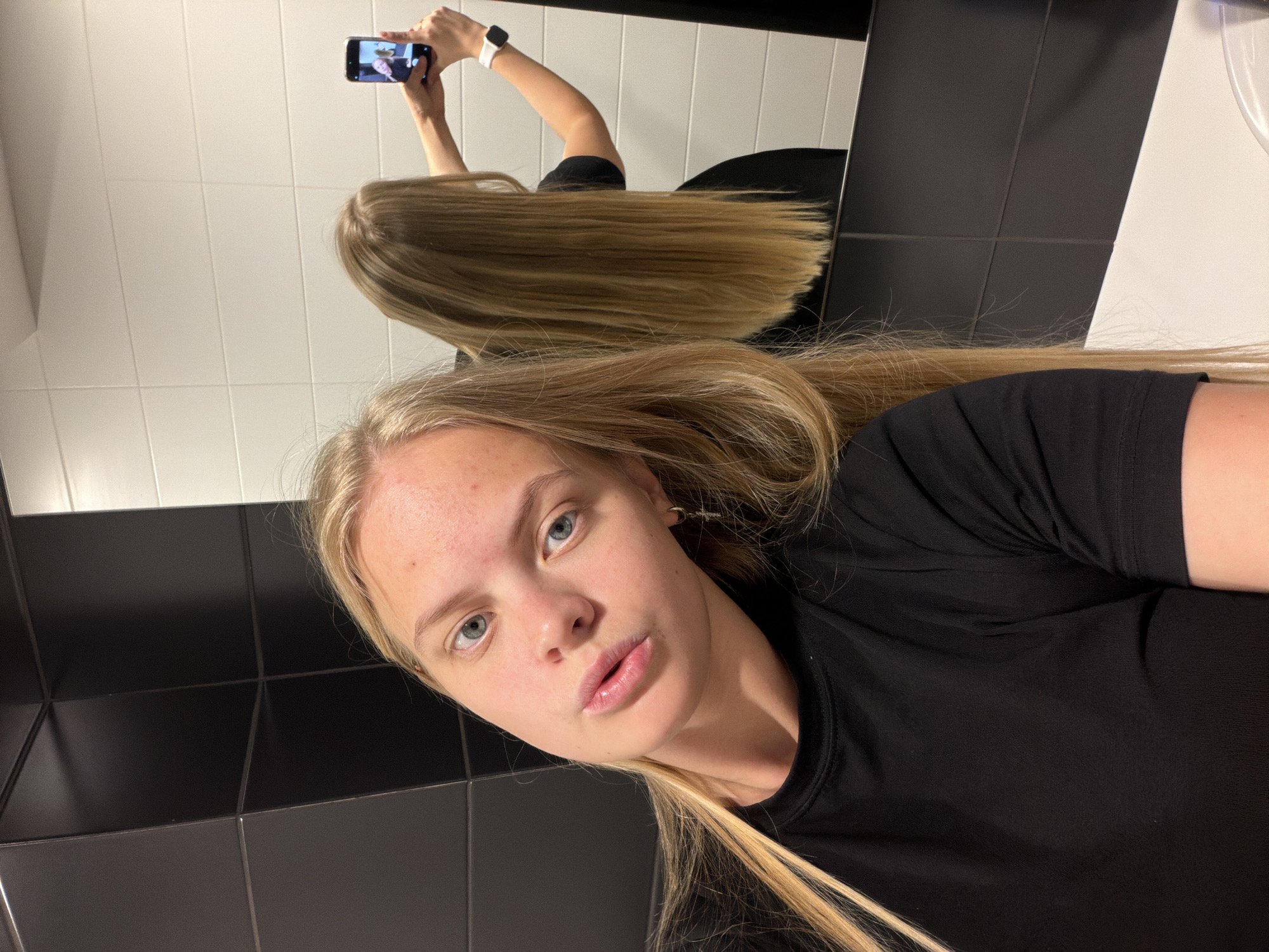 A young woman with long, straight blonde hair and light blue eyes takes a mirror selfie in an indoor setting. She has fair skin with some minor blemishes on her forehead and is looking directly at the camera with slightly parted, pinkish lips. She wears a plain black short-sleeved t-shirt and a simple earring in her left ear.
The background consists of tiled walls. The wall to her right and beneath her face is made of large black square tiles with thin dark grout lines. In the mirror, behind her reflection, white rectangular tiles with dark grout are visible, suggesting a distinct section or wall.
Her reflection is prominent in the mirror, showing her holding a dark-colored smartphone overhead with both hands to capture the image. A white smartwatch or fitness band is visible on her left wrist in the reflection. The phone screen displays some content, likely another image or video, but no specific text is legible. Her long blonde hair flows down her back and is also visible in the reflection.
The image captures a casual moment indoors, likely in a restroom or changing room. The lighting appears artificial. The overall composition is a diagonal self-portrait, giving it a dynamic feel. No clear text is visible in the image. The location context for this scene is Izhevsk, Russia.