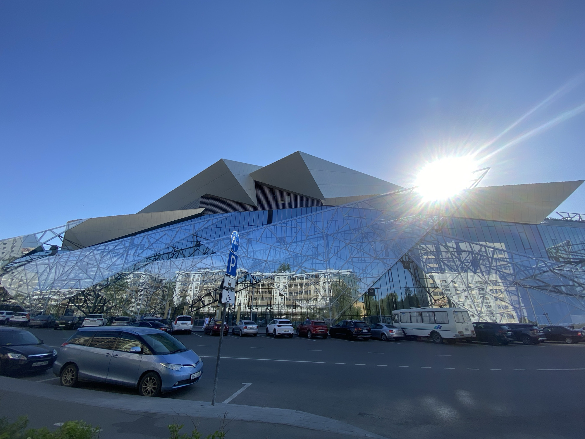 The image depicts a modern architectural structure in Kazan, Russia, under a clear blue sky with bright sunlight. The building features a complex facade of glass and angular shapes, with reflections of surrounding buildings visible in the glass. In front of the building is a parking area filled with various cars and a white van. A blue van is parked in the foreground. A parking sign with a bicycle and handicap symbol stands beside it. The pavement and a small patch of greenery are also visible in the foreground.