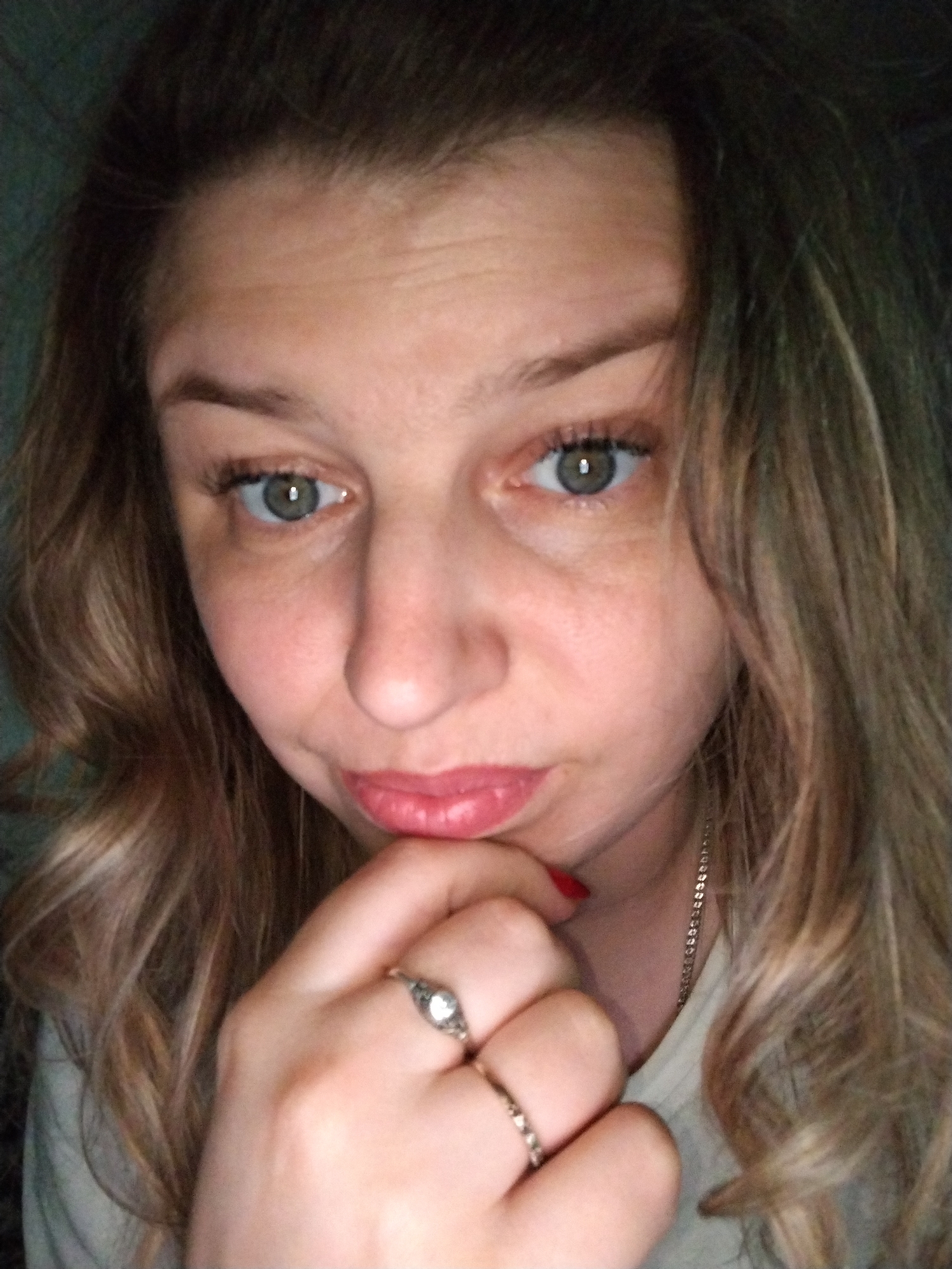 A fair-skinned woman with light brown wavy hair is pictured in a close-up selfie. She is wearing a light-colored top and a thin silver necklace. Two silver rings are visible on her hand, which is posed with her fingers touching her chin. Her eyes are light-colored, and she is looking directly at the camera with a slight frown. The background is dimly lit and appears to be an indoor space. The location in the city of Rusanovo, Russia is not discernible from the visual information provided.