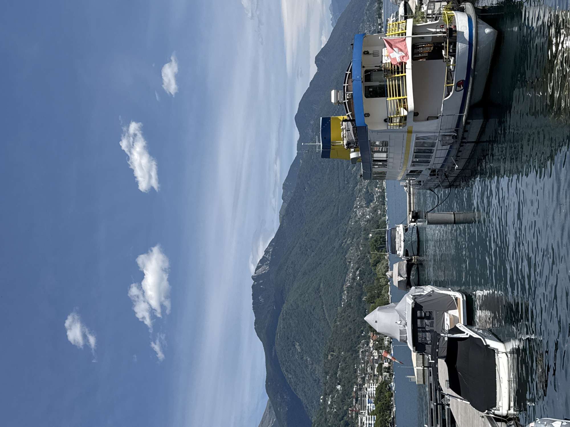 The image depicts a scenic view of the Locarno waterfront in Switzerland on a clear, sunny day with scattered clouds. In the foreground, several boats are docked, including a large white and blue passenger ferry with a Swiss flag prominently displayed on its stern. Adjacent to the ferry, smaller pleasure boats are moored, some covered with protective tarps. The water is calm, reflecting the surrounding scenery.
In the mid-ground, the city of Locarno is visible, with buildings nestled along the shoreline and extending up the slopes of lush green mountains. These mountains form a dramatic backdrop to the scene, with their forested slopes reaching towards the sky. The sky itself is a vibrant blue, punctuated by fluffy white clouds. The overall impression is one of a peaceful and picturesque lakeside setting.