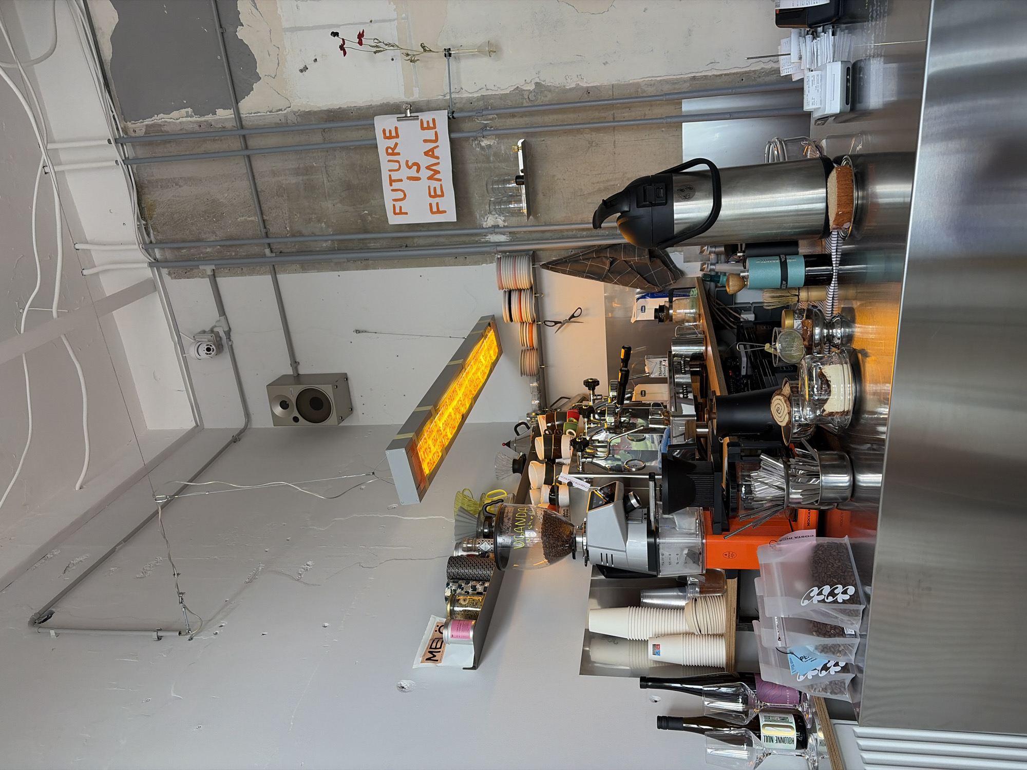 This indoor image captures a coffee preparation area, likely within a modern cafe or bar in Belgrade, Serbia. No people are visible. The scene is dominated by a stainless steel counter on the right, laden with various coffee-making equipment.
On the counter, a prominent coffee grinder with a clear hopper holds coffee beans, the hopper clearly marked "UGANDA". An espresso machine, various metal coffee tools like whisks and stirrers, and stacks of disposable white paper cups are arranged around it. Several transparent bags of coffee beans are visible, along with multiple bottles, one of which clearly reads "KOLONNE NULL". A large silver insulated dispenser stands at the end of the counter. Receipts are clipped to an apparatus above the counter, reflecting against the metallic surface.
The walls are painted a light color, contrasting with the exposed grey electrical conduits that run horizontally across them. Mounted on the wall, above the counter, is a rectangular industrial-style heater or lamp emitting a bright orange glow. Repetitive text, including "GRATIS GRATIS GRATIS", is visible on its surface. To the left, a grey speaker is mounted high on the wall.
The ceiling above is raw concrete, featuring a network of grey and white exposed pipes and conduits, reinforcing an industrial aesthetic. A white sign with bold orange capital letters reads "FUTURE IS FEMALE", clipped to one of the pipes. Small decorative elements resembling dried red flowers or berries are attached to the ceiling near this sign. A white security camera is also mounted on the wall. A small item on a shelf below the heater has the letters "ME" visible.