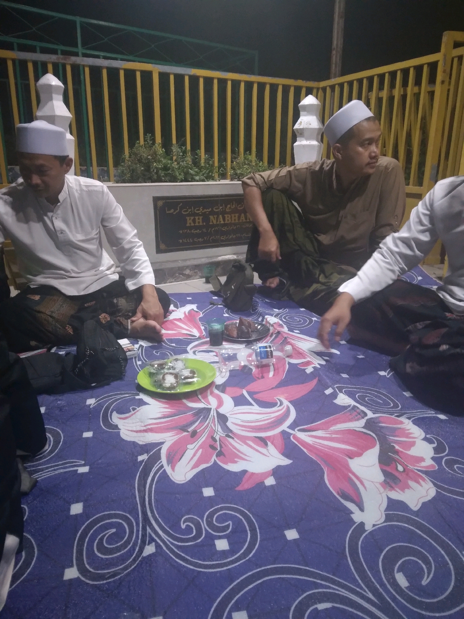 Three adult males are seated on a blue patterned mat in what appears to be a cemetery in Batam, Indonesia. The man on the left wears a white peci hat and a white long-sleeved shirt with a black sarong. The man in the center wears a brown long-sleeved shirt and a dark green sarong. The man on the right, partially visible, wears a white long-sleeved shirt and a dark sarong. In front of the men are plates of food, a bottle of water, and a glass. Behind them is a gravestone with Arabic inscriptions and a yellow metal fence. The lighting suggests it is nighttime.