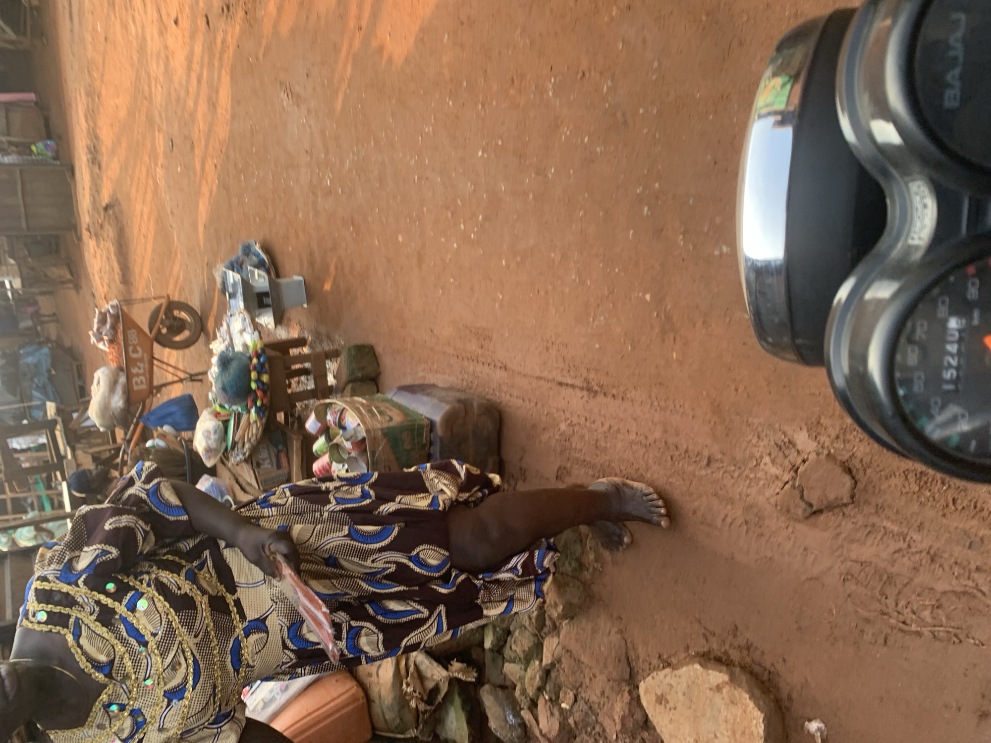 A woman in a brown and blue patterned dress with gold embellishments is standing barefoot on a dirt road in Shagamu, Nigeria. She is holding a red fan. To her left, there are various items for sale, including what appears to be canned goods in a basket, a scale, and nets. In the background, there are market stalls and a wheelbarrow filled with goods. The ground is reddish-brown dirt, and there are some scattered stones and debris. The scene suggests an outdoor market environment. A motorcycle's dashboard is visible in the foreground.