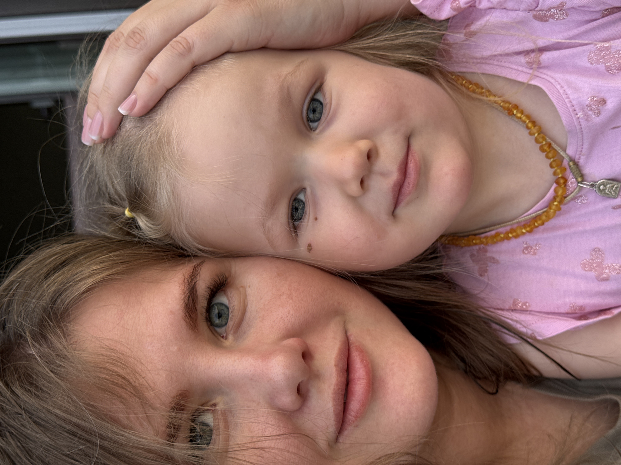 A close-up shot depicts a woman and a young child, seemingly intertwined in a familial embrace. The woman, with light brown hair and blue eyes, is positioned at the bottom of the frame, while the child, sporting blonde hair and blue eyes as well, rests her head atop the woman’s, with a hand gently placed on her head. The child is wearing a pink shirt decorated with butterfly patterns and an amber beaded necklace with a silver pendant. They are likely located in the city of Ферма, Russia.