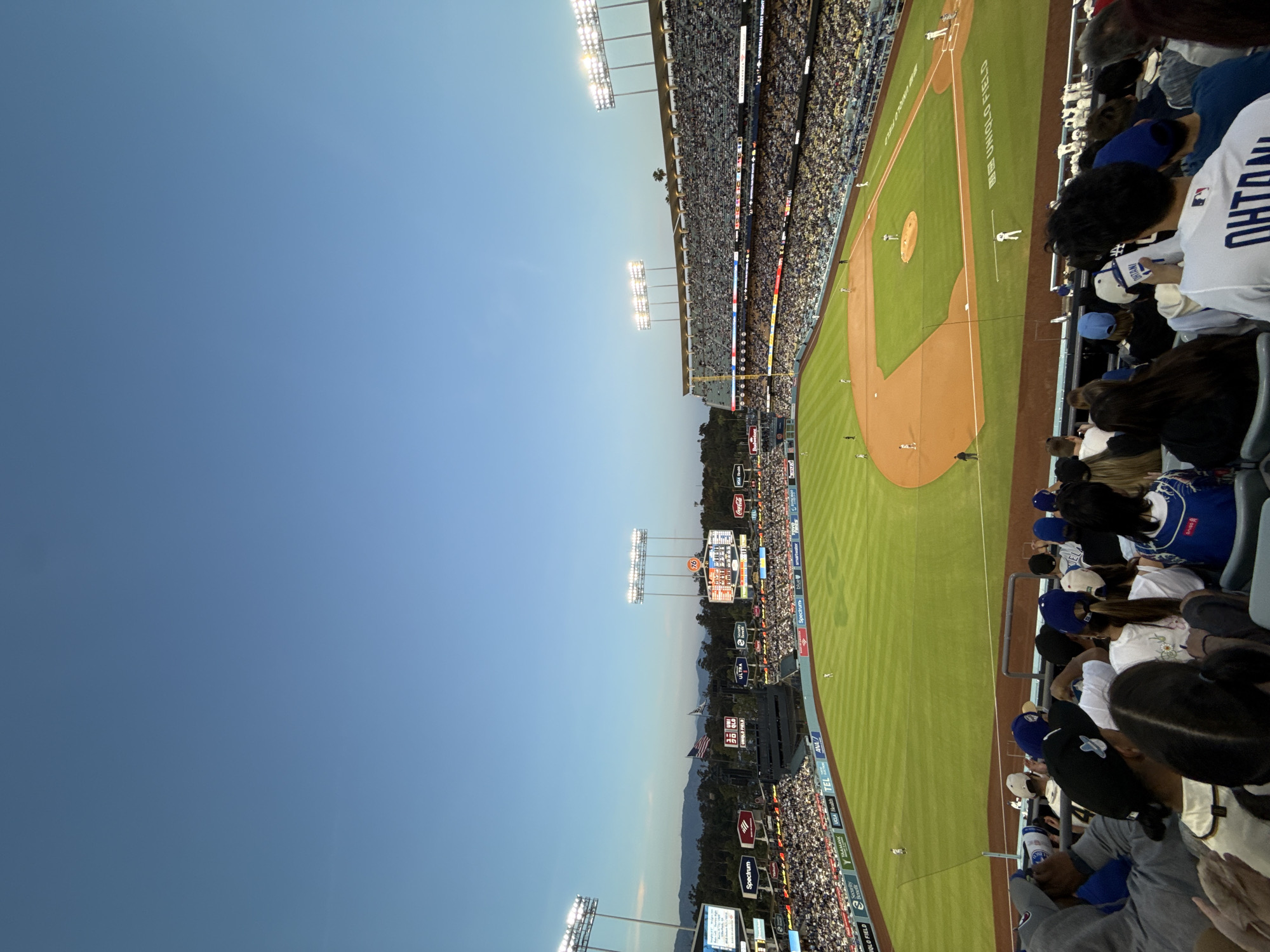 This image shows a baseball game in progress at Dodger Stadium in Los Angeles, USA. The stadium is full of fans and the game appears to be during the early evening, as indicated by the natural light and stadium lights. The crowd is predominantly wearing blue, signifying support for the Los Angeles Dodgers. Banners and advertisements are visible along the outfield wall and above the stands.