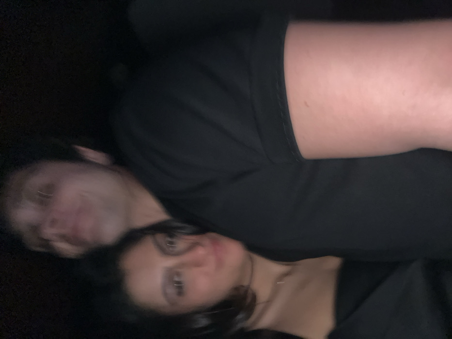 The image captures a close-up, diagonally oriented shot of two individuals, likely young adults, in a very dimly lit environment. Both subjects exhibit significant motion blur, especially on their faces, suggesting movement during a long exposure or a quick snapshot in low light.
In the foreground, positioned towards the lower right of the frame, is a person with dark hair, which appears to be long and flowing around their head. Their face, though blurred, is turned towards the camera, and they seem to be looking directly into the lens. Visible features include a nose piercing and possibly other facial piercings. They are wearing dark clothing, likely black, which covers their upper body.
Slightly behind and to the left of this person, partially obscured, is another individual. This person appears to have lighter hair and their face is more blurred and less distinct, but they are also looking generally in the direction of the camera. The two individuals are positioned very closely, suggesting intimacy or companionship, possibly lying down side-by-side or one resting on the other.
The upper right portion of the image shows a bare arm or leg with fair skin tone, belonging to one of the individuals, contrasting with the dark fabric of their clothing. The clothing worn by both individuals is predominantly black or a very dark shade, blending seamlessly with the almost entirely black background.
The overall setting is extremely dark, offering no discernible details about the surroundings beyond the subjects themselves and their clothing. The dim lighting and blur create a soft, somewhat mysterious atmosphere. There is no visible text in the image.