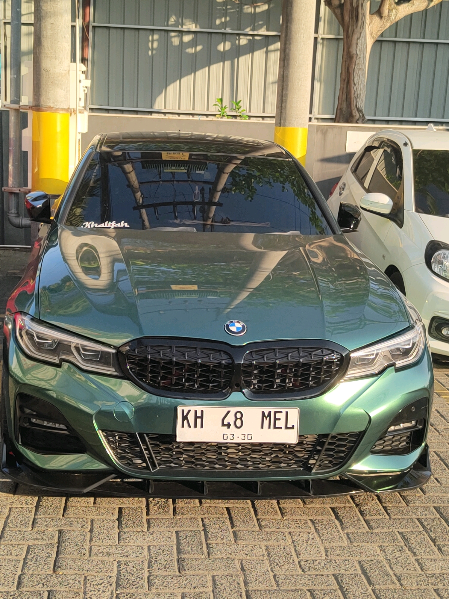 An eye-level, full front shot captures a striking green BMW sedan parked on a patterned brick surface. The car is the central subject, its metallic paint gleaming under natural light. Its prominent black grille and sleek headlights are clearly visible, as is a black front splitter. A license plate reading "KH 48 MEL" is mounted centrally below the grille. A white sticker with the word "Khalifah" is affixed to the upper windshield.
In the background, a light-colored hatchback is parked to the right of the BMW. Beyond the vehicles, a corrugated metal wall forms the backdrop, with a large tree trunk visible to the right. A concrete pillar with a yellow band is situated to the left of the BMW, and a smaller green plant grows near the base of the metal wall. The overall environment appears to be an outdoor parking area, likely within a commercial or residential complex in Banjarmasin, Indonesia. The light suggests it is daytime, possibly late afternoon due to the long shadows. No people are present in the frame.