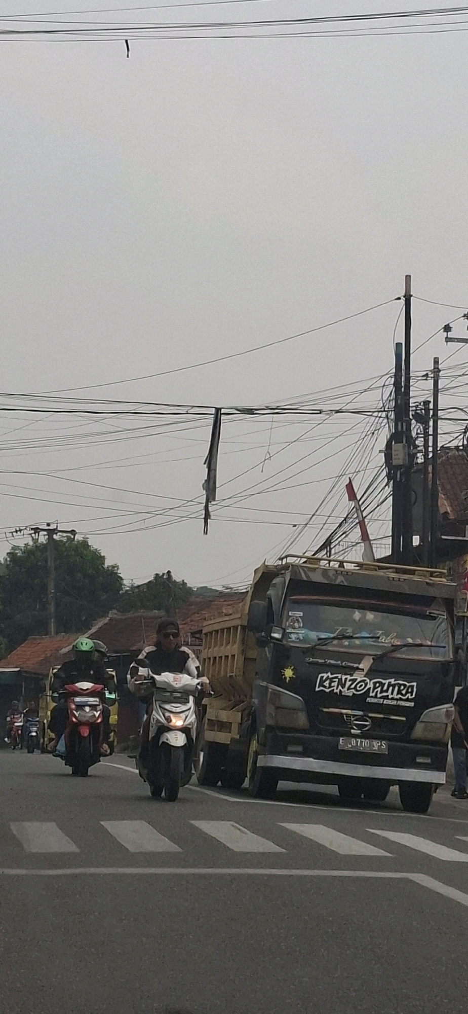 This is a vertical, eye-level shot of a street scene, likely in Sirnagalih, Indonesia, characterized by a slightly overcast sky and a dense network of overhead electrical wires. The foreground features a pedestrian crossing marked by white stripes on asphalt. On the street, a black dump truck with a wooden bed is prominent, bearing the text "KENZO PUTRA" and a license plate reading "E 0770 NPS". To the left of the truck, two individuals on motorcycles are visible. The closest motorcycle, a white scooter, is ridden by a person wearing sunglasses and a black and white jacket. Behind them, a red motorcycle is ridden by a person wearing a green helmet. Further down the street, other vehicles and buildings with red tile roofs are visible. A tattered black fabric is hanging from one of the overhead wires, adding a touch of disarray to the scene. The overall impression is one of an active, possibly congested, urban or semi-urban roadway.