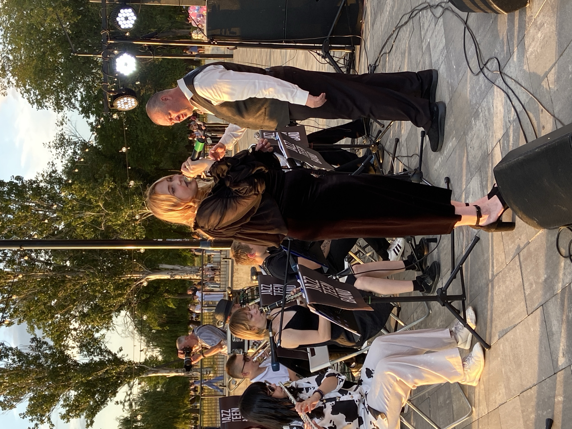 A medium shot captures an outdoor jazz performance at dusk. In the foreground, a young woman with blonde hair, dressed in a black velvet dress, sings into a microphone while standing to the left of a bald man in a white shirt and dark vest. Behind them, a band of young musicians is seated, playing instruments including a flute and a clarinet. Music stands with "Jazz Teen Band" printed on them are visible. In the background, trees, string lights, and a crowd of people are visible. A photographer is seen in the middle ground, taking a picture of the event. The ground is paved with square tiles. The lighting suggests it is late afternoon or early evening.