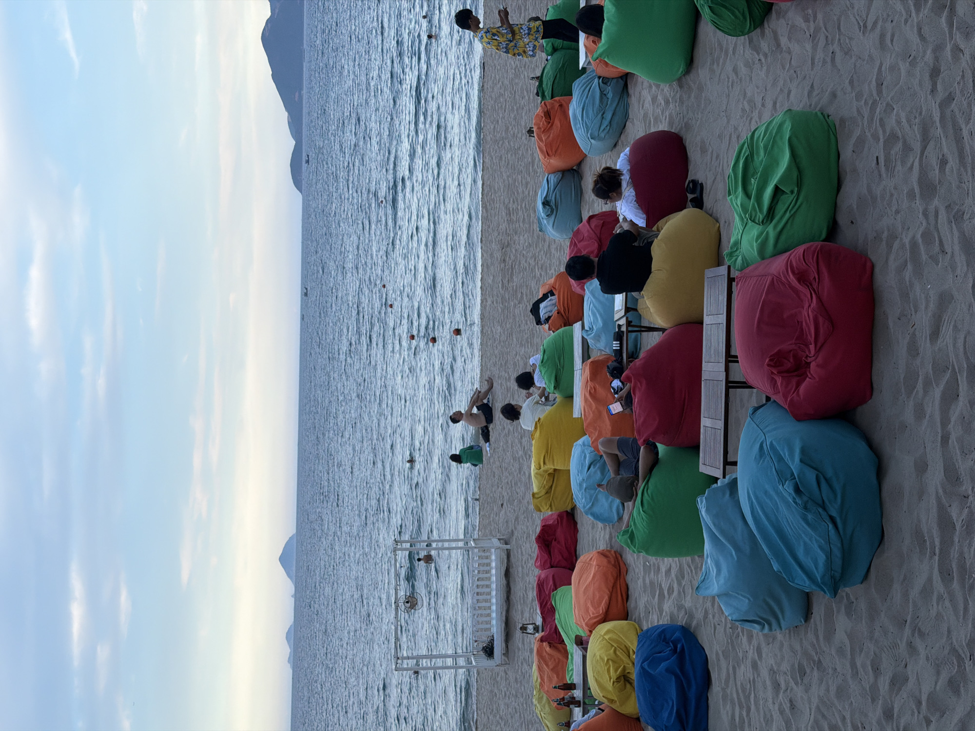 The image depicts a beachside seating area in Nha Trang, Vietnam, with numerous large, colorful beanbag chairs scattered across the sand. The ocean stretches out in the background under a cloudy, pale blue sky, with a few distant islands visible on the horizon. Several people are seated on the beanbags, some facing the water and others engaged in conversation. In the middle ground, a white, open-sided canopy structure is set up on the sand, with a fan mounted inside. A few individuals are in the shallow water, swimming or wading. To the right, a person in a vibrant yellow floral shirt is looking at their phone. The overall scene conveys a relaxed, leisurely atmosphere typical of a beach resort.
