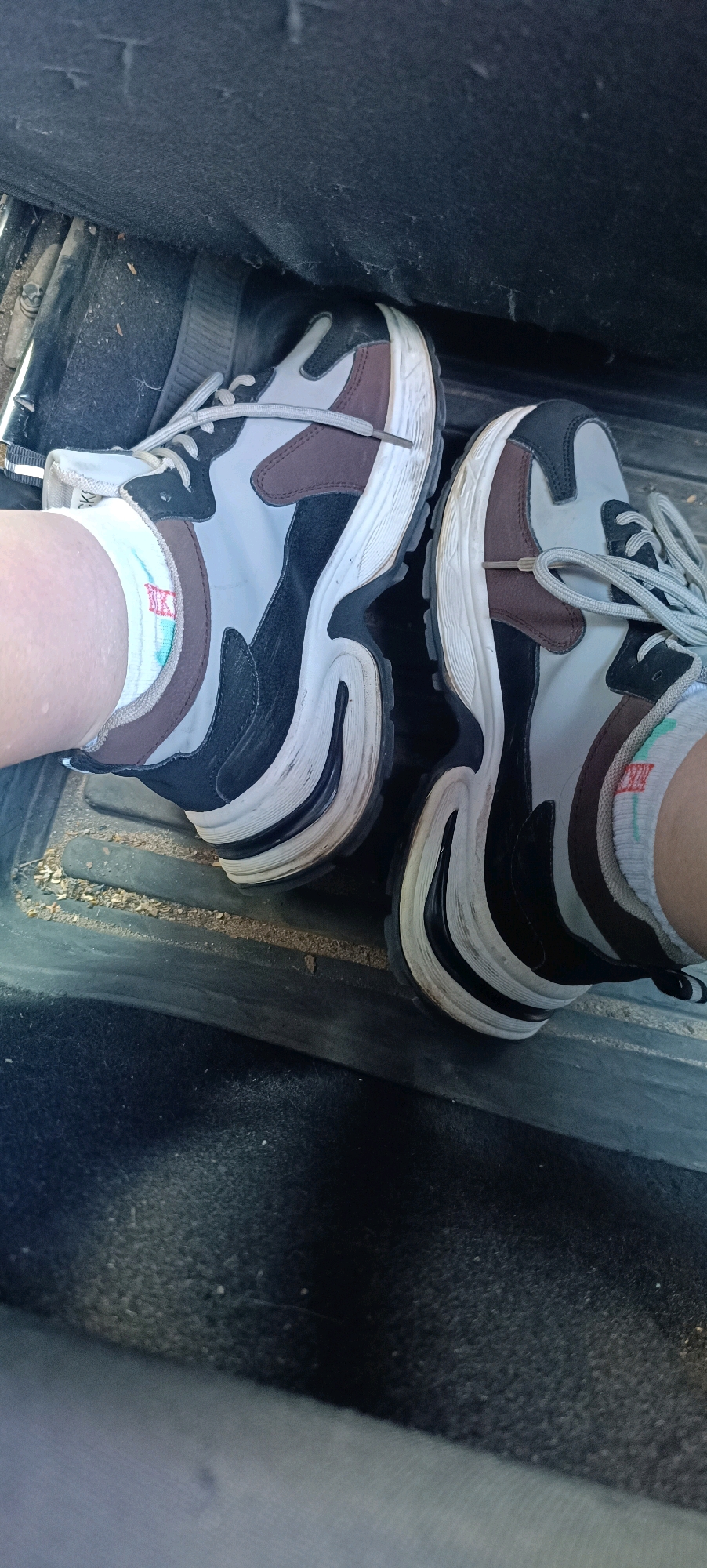 The image shows a person's feet wearing athletic shoes inside a vehicle. The shoes are multi-colored, with sections of grey, black, and brown, and have thick white soles. The person is wearing white socks with colored stripes. The vehicle's interior appears to be a floor mat area, with some debris on the mat. The background is dark and out of focus. There is no discernible information in the image that could place the location in the city of Perm, Russia.