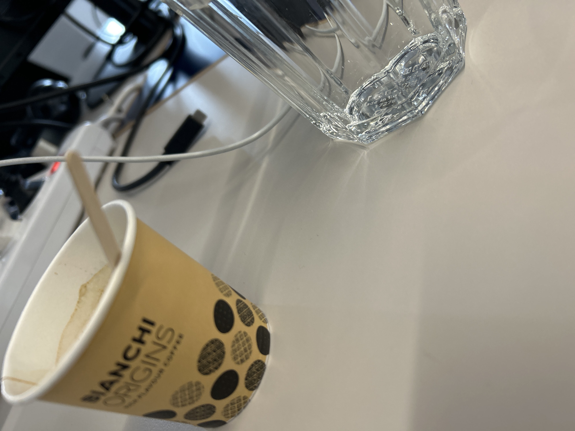 The image presents a slightly oblique close-up shot of a desk scene. A partially consumed cup of coffee, branded "BIANCHI ORIGINS", rests on the desk surface alongside a clear, faceted glass. A small wooden stirrer is inside the coffee cup. Cables and a power strip are visible in the background, suggesting a workspace setting.
Without additional information, pinpointing the exact location in Sofia, Bulgaria, is impossible. However, the style of the coffee cup and glass doesn't reveal specifics about the building.