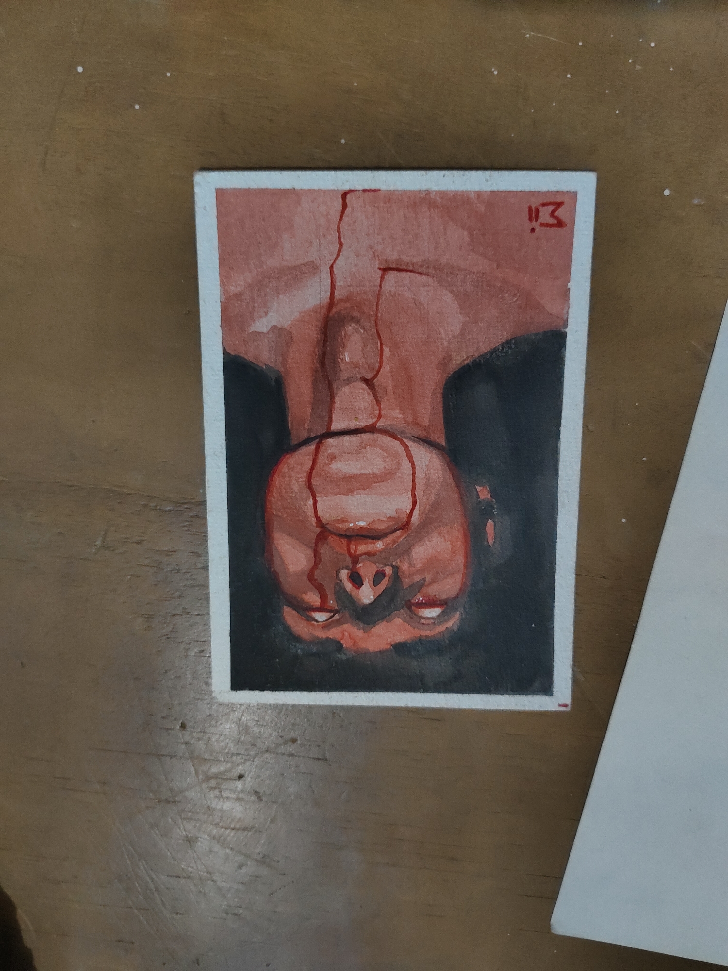 A small, rectangular watercolor painting is depicted centrally on a light brown wooden surface. The painting features an inverted human figure, visible from the upper chest to the head. The skin tone is rendered in shades of reddish-brown and pink, with prominent red streaks, consistent with blood, flowing from the facial area (specifically, appearing to emanate from the nose/mouth region) downwards across the neck and chest. The figure's eyes are open and show a direct gaze. The background within the painting is a solid, dark tone. A red mark resembling "!!3" or "!!E" is present in the upper right corner of the painting.
The wooden surface underneath the painting exhibits visible wood grain, numerous fine scratches, and a sparse scattering of small white particulate matter. To the immediate right of the painting, a flat, off-white surface, possibly paper or cardboard, is partially visible. The scene is illuminated by diffuse, overhead lighting. The image contains no discernible features that indicate a specific geographic location, including Davao City, Philippines.