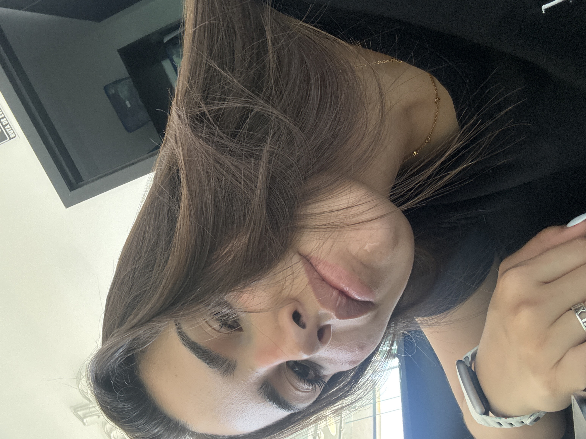 A close-up, rotated image captures a young woman from a low, upward angle, with her long, wavy dark brown hair framing her face. She has prominent, well-groomed eyebrows, defined eyes with eyeliner and mascara, and slightly parted lips with what appears to be light-colored lipstick. Her expression is neutral.
She wears a delicate gold chain necklace around her neck, partially visible beneath her hair, and a black top. Her left hand is visible, featuring a smartwatch with a light-colored, patterned band on her wrist and a silver ring adorned with Roman numerals (specifically "XII" and "II") on her ring finger. Her fingernails are painted a light shade, possibly white or light blue.
The background suggests an indoor setting. To the upper left, there's a dark, reflective rectangular surface, possibly a screen or window, showing a faint reflection of what appears to be a light-colored object or another screen. Adjacent to this, a white sign with black text is partially visible, reading "RUTA DE EV...". The overall context is San Bernardino Tlaxcalancingo, Mexico. The woman appears to be posing for a selfie or a close-up portrait.
