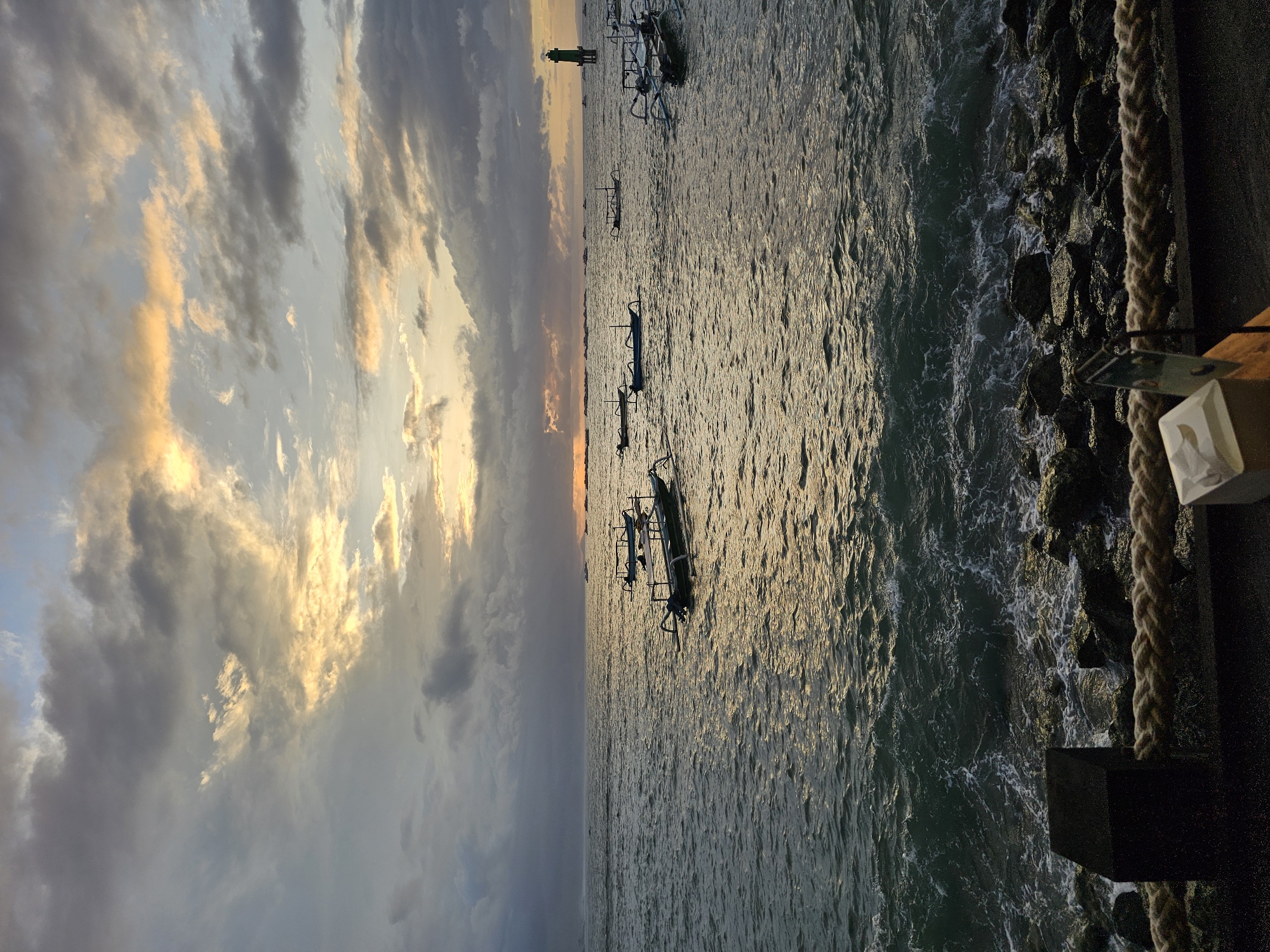 Captured at sunrise or sunset, the scene features a coastline in Denpasar, Indonesia, with several traditional outrigger boats, known as "jukung," resting on the water. A green navigational marker stands distantly in the water. Dark rocks protect the shoreline. The sky above exhibits mixed cloud cover, with golden light filtering through the clouds near the horizon. A dock edge is on the right side of the image with rope. A tissue box rests on the dock.