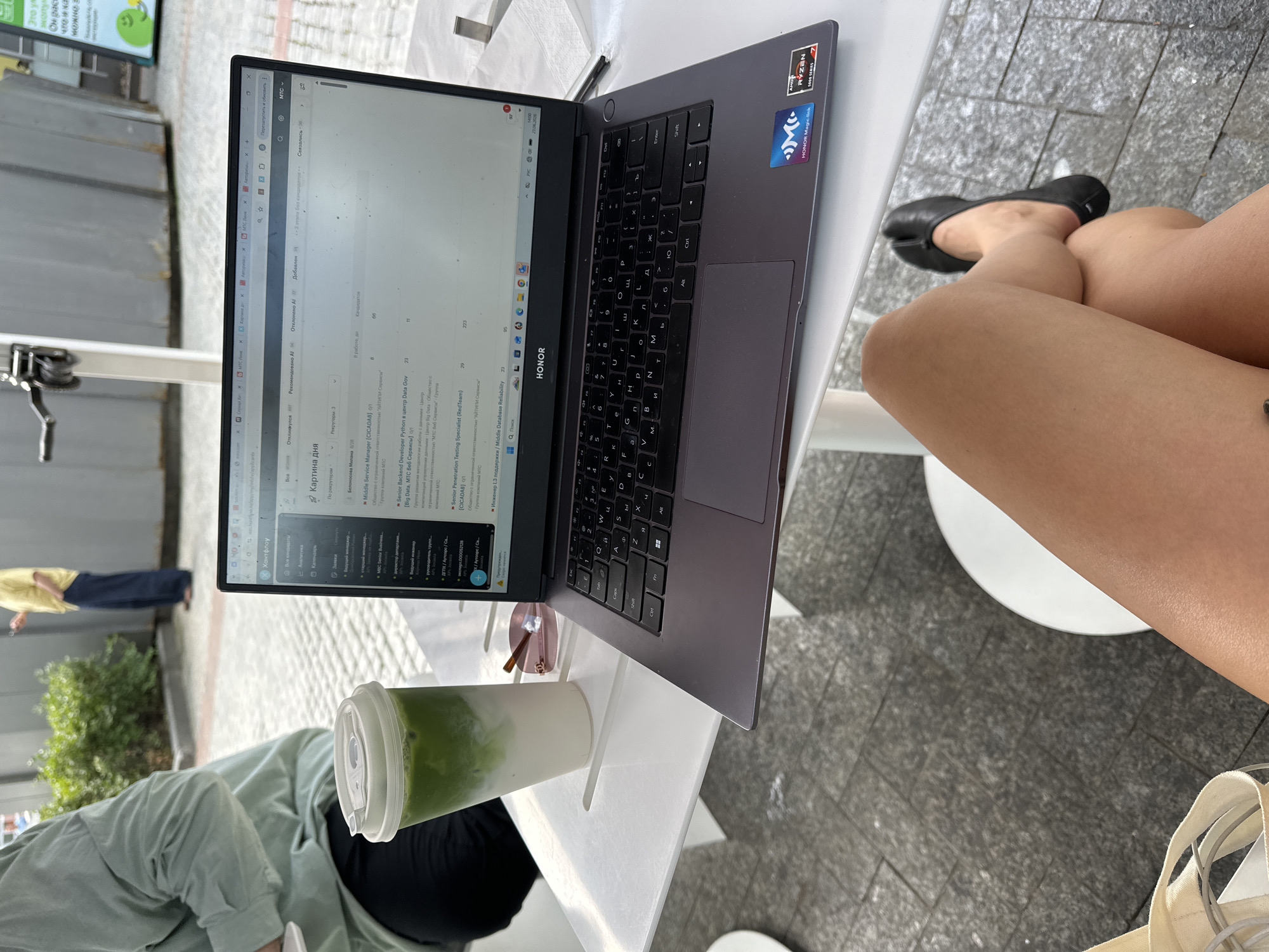 A high-angle, vertical shot captures a person working on a laptop at an outdoor table. The laptop, prominently displayed, shows a webpage with text in Russian, likely a job board or recruitment platform, with job titles like "Senior Backend Developer Python" and "Senior Penetration Testing Specialist." The laptop's brand is visible as HONOR, and it has AMD Ryzen 5000 Series branding.
Beside the laptop is a clear plastic cup filled with a layered green and white beverage, possibly a matcha latte. The person's legs and feet are visible, wearing black ballet flats. They are seated on a light-colored chair, and a canvas bag rests near their feet.
The setting appears to be an outdoor seating area with a gray tiled ground and a white table. In the background, a person in a yellow shirt and dark pants walks past, and some greenery is visible. The overall impression is of someone working remotely or studying outdoors, perhaps during the daytime given the bright lighting, though no specific weather is apparent. The time on the laptop screen reads "14:00 23.06.2026."