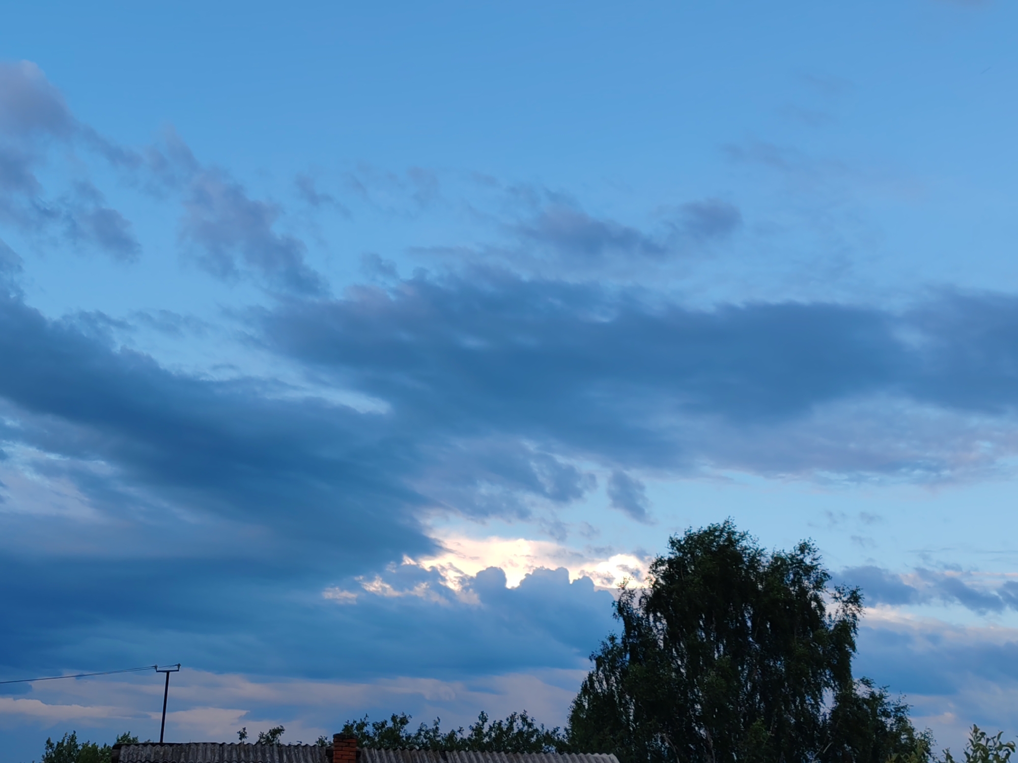 The image depicts a cloudy sky with hues of blue and grey, illuminated by a bright patch of sunlight breaking through the clouds. In the foreground, the top of a roof made of corrugated material is visible, with a chimney and some trees lining the horizon. A dark tree with dense foliage is prominently featured on the right side of the image. A thin pole with wires stretches across the lower left portion of the frame, adding a man-made element to the natural landscape. The scene suggests an outdoor setting, possibly in a rural or suburban area, with a focus on the atmospheric conditions of the sky. The location is specified as Чухлэм, Russia.