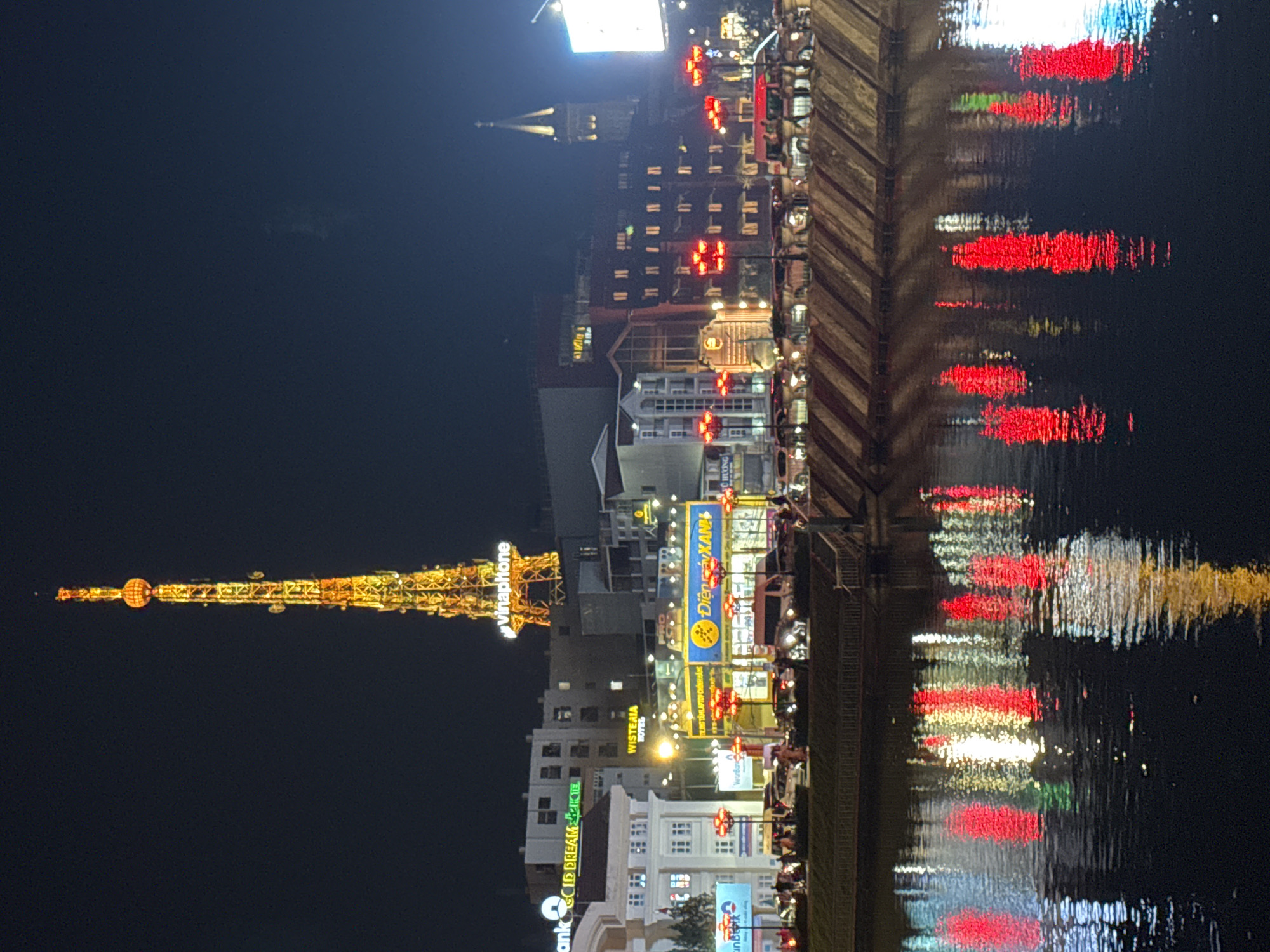 This media file captures a nighttime scene in Da Lat, Vietnam, likely near a body of water given the prominent reflections. The image is vertically oriented.
The main subjects include a tall, illuminated tower resembling a replica of the Eiffel Tower, adorned with golden lights. Nearby, buildings of various heights are visible, some lit with numerous windows, while others feature illuminated signs. Among the visible text on these signs are "vinaphone," "WISTERIA HOTEL," "CID DREAM," and "Điện gáy XAN." The reflections of the lights from the buildings and the tower are clearly visible on the dark water in the foreground, creating a vibrant display of red, white, and yellow streaks.
The scene is set at night, as indicated by the darkness of the sky and the illuminated structures. The overall atmosphere appears lively, suggested by the abundance of lights and the bustling cityscape implied by the presence of hotels and businesses. The weather is not explicitly depicted, but the reflections suggest a calm water surface. The scene evokes a sense of urban nighttime charm, typical of a tourist destination like Da Lat.