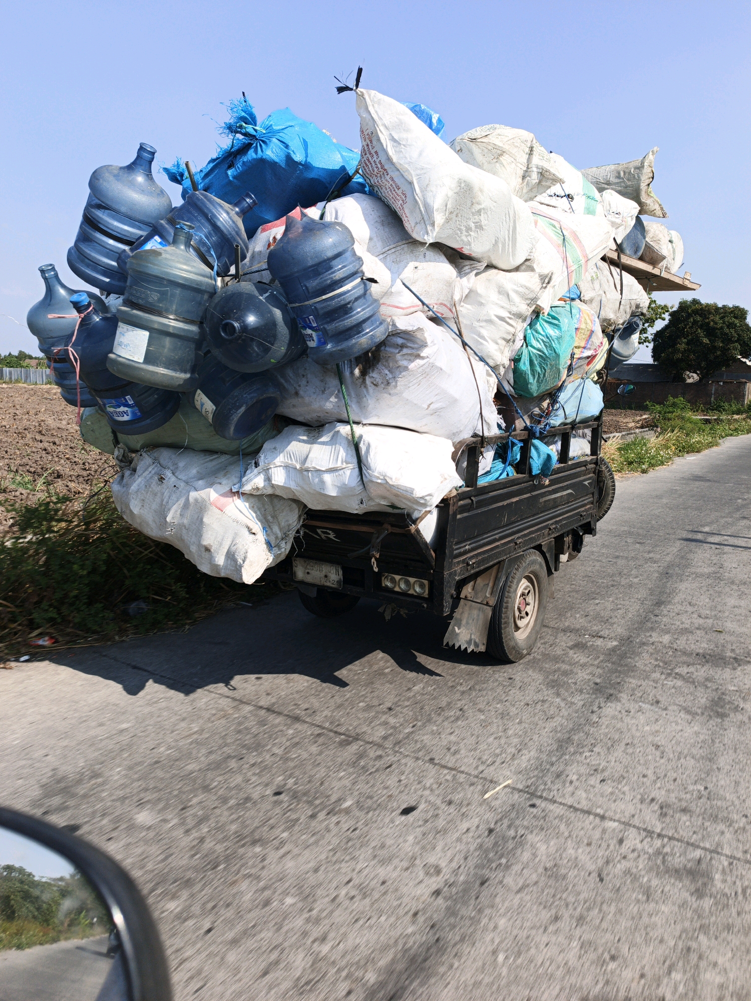 The image shows a black, three-wheeled truck overloaded with various items, including large blue water jugs and white sacks, driving on a paved road. The truck is positioned in the center of the frame, slightly angled away from the viewer. The background consists of a clear blue sky, a field with tilled soil, and some vegetation. On the right side of the road, there is a cluster of trees and buildings, indicating a semi-rural or suburban environment. The perspective suggests the photo was taken from another vehicle moving in the same direction. The overall scene depicts a common sight of overloaded vehicles transporting goods, likely for recycling or distribution, in a sunny, outdoor setting. The location is identified as the city of Kejagan, Indonesia.