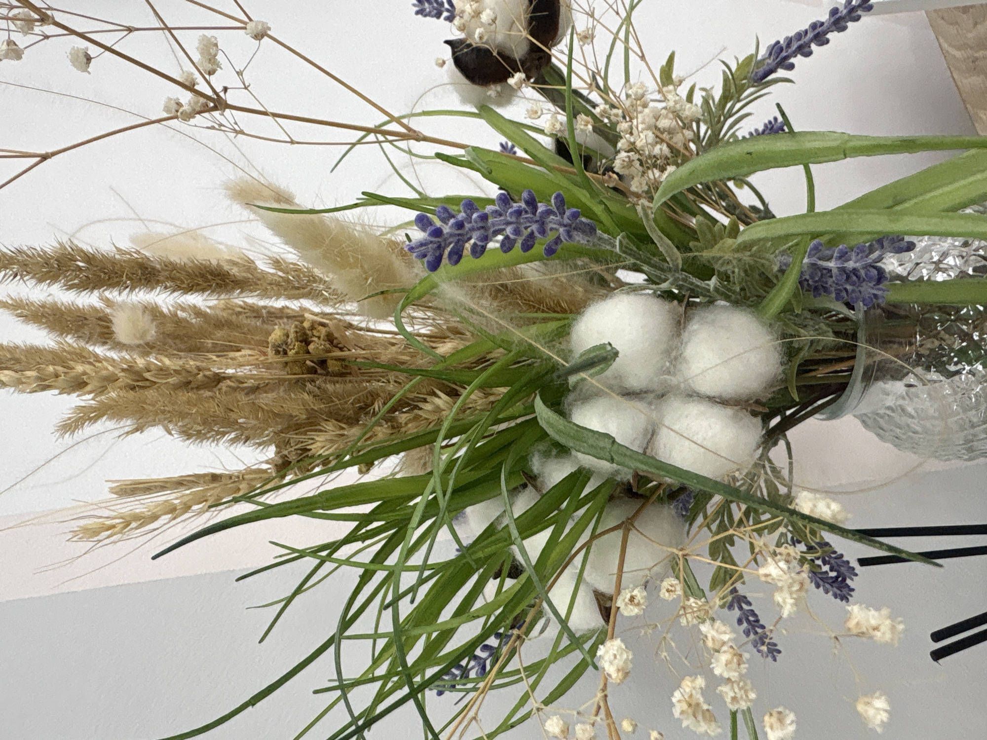 The image shows a floral arrangement with dried grasses, cotton bolls, lavender, and baby's breath. The bouquet appears to be in a clear glass vase. The background is a plain white wall. No evidence indicates this image was taken in Perm, Russia.