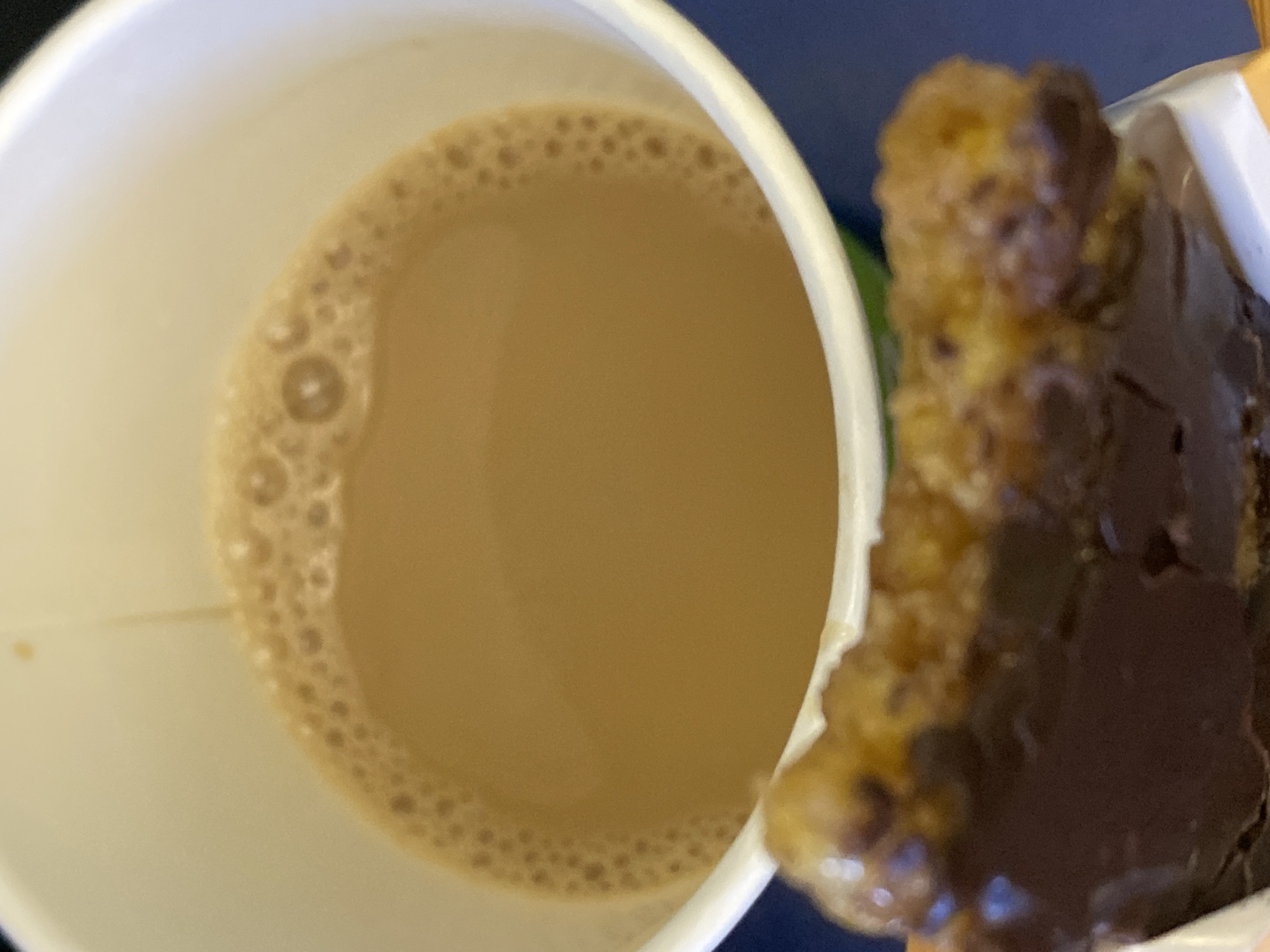 The image shows a close-up of a white paper cup filled with a light brown beverage, likely coffee or tea with milk, topped with a layer of small bubbles. To the right of the cup is a dark brown, rectangular-shaped food item, possibly a cookie or brownie, with visible chocolate icing and a textured, golden-brown base. The background is a dark blue, suggesting an indoor setting or a dark environment. The lighting is soft, and the focus is primarily on the cup and the food item. There are no people visible, and no text is apparent in the image. The overall impression is a simple depiction of a drink and a sweet treat, possibly for a snack or breakfast. The context of Ak-Jol, Kyrgyzstan, does not provide specific visual cues within the image itself.