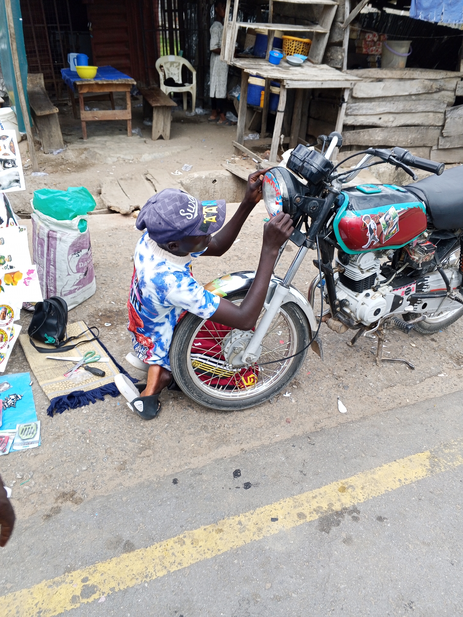 A person is kneeling on the pavement next to a motorcycle, appearing to be making adjustments to the front headlight in Zango-ndaji, Nigeria. The person is wearing a patterned blue and white shirt, red shorts, a baseball cap, and slippers. The motorcycle is parked on the side of a paved road marked with a yellow line. In the vicinity of the motorcycle are a sack, a small rug with accessories, and other items. The background includes a ramshackle wooden structure with various objects on shelves and tables.