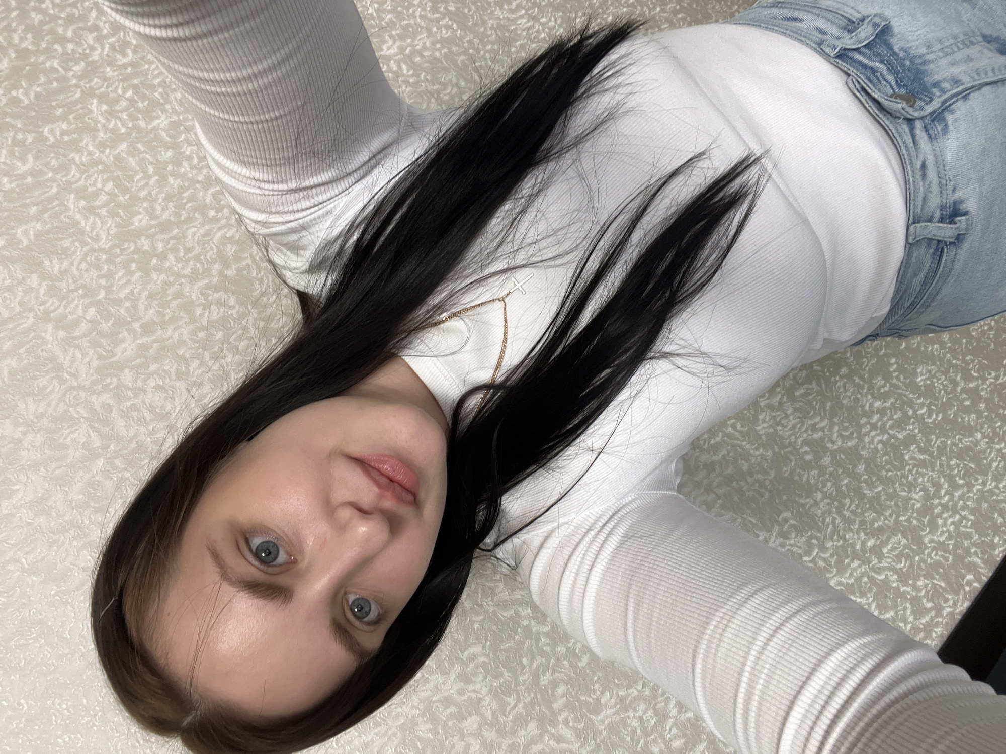 A woman with fair skin, blue eyes, and dark brown hair is depicted in a selfie-style photograph. She is wearing a white long-sleeved ribbed shirt and blue jeans. A gold necklace with a cross pendant is visible around her neck. The background appears to be a patterned surface. The image appears to be taken indoors, but the exact location in Noviy, Russia, is not discernible.