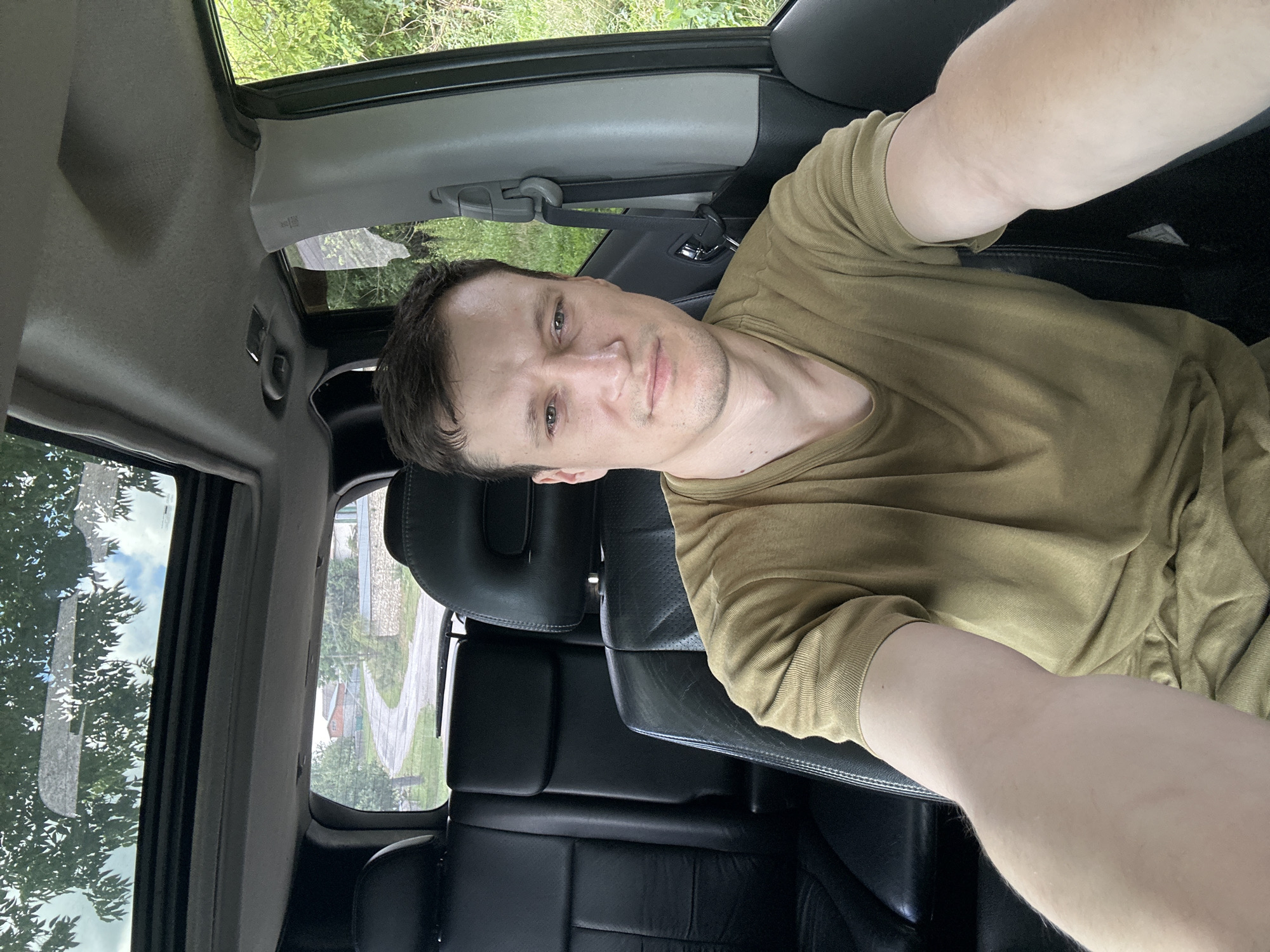 A man with short brown hair and light skin is sitting in the driver's seat of a car, taking a selfie. He is wearing a plain olive green t-shirt and has a neutral expression. The car has black leather seats and a sunroof, which is open, allowing light to filter in. Through the open sunroof and the windshield, a rural landscape is visible. It appears to be daytime, with trees and houses in the background, suggesting a location outside of a major city. The image is taken from a slightly elevated angle.