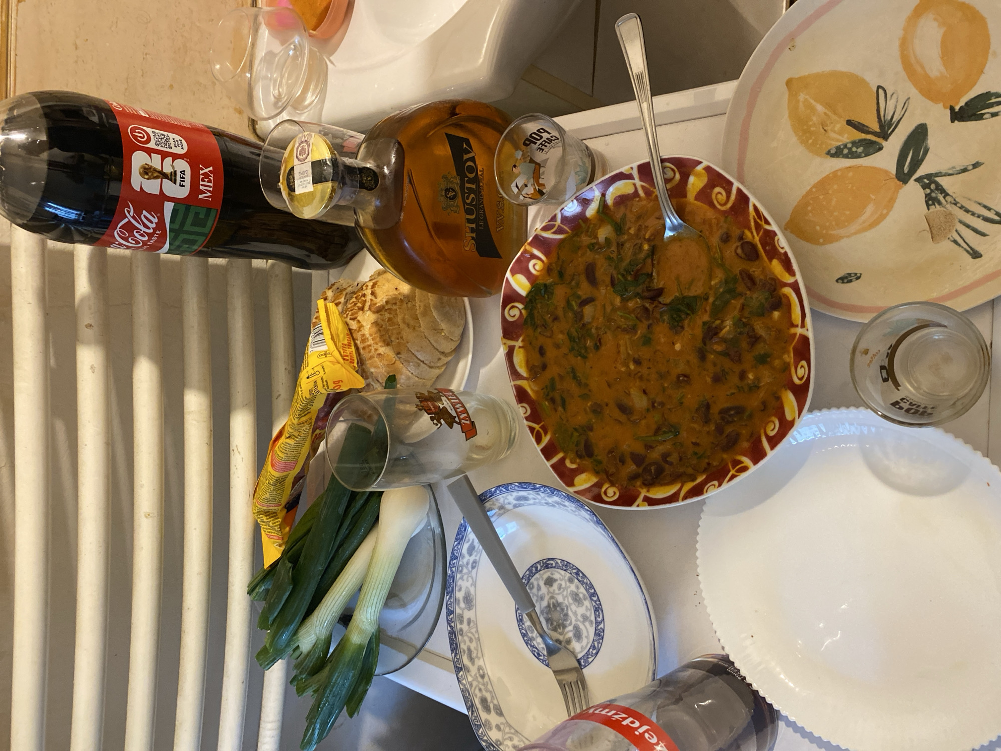 This is an overhead, close-up, indoor, eye-level shot of a table laden with food and drinks, possibly at a home in Szczecin, Poland. A large bottle of Coca-Cola, labeled "MEX" and featuring FIFA branding, stands prominently. Next to it, a bottle of Shustov cognac is visible, along with a small shot glass. A plate of what appears to be chili or stew with green vegetables and beans is centrally located, accompanied by a fork. Other items on the table include green onions, sliced bread, a plate with a blue and white floral pattern, a plain white plate, and a decorative plate with painted lemons. The background shows a white radiator. The overall impression is of a casual meal or gathering.