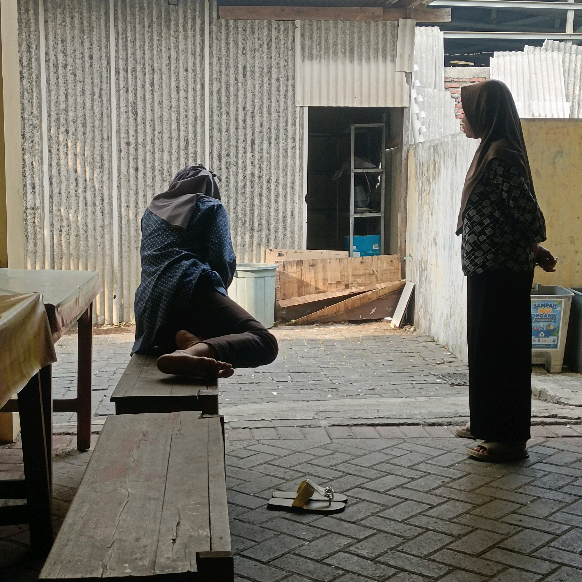 Two women are in an outdoor setting, likely a backyard or alleyway in Surabaya, Indonesia. The woman on the left is sitting on a wooden bench, facing away from the camera, with her head down, wearing a blue patterned top and dark pants, and a grey headscarf. Her feet are visible and bare. The woman on the right is standing, facing towards the left, wearing a dark patterned top and dark pants, and a brown headscarf. She is looking towards the seated woman.
The environment appears to be utilitarian, with corrugated metal walls in the background, a doorway leading to an interior space with shelves, and piles of wooden planks. A light-colored trash bin with Indonesian text "SAMPAH ORGANIK" (Organic Waste) is visible to the right. The ground is paved with gray bricks laid in a herringbone pattern. A table with a light-colored tablecloth is present on the left side of the frame, and a pair of light-colored flip-flops with a decorative element are on the ground in the foreground. The lighting suggests it is daytime, with some shadows cast on the ground.