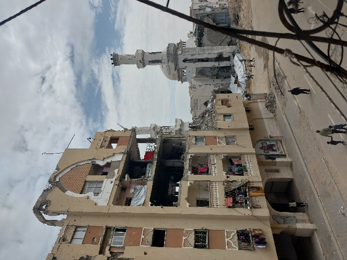 The image depicts a street scene in Khan Yunis, Palestinian Territory, characterized by significant structural damage under a partly cloudy sky. In the foreground, a heavily damaged multi-story residential building dominates the left side. Its beige facade shows large sections of missing plaster and concrete, exposing interior rooms, rebar, and brickwork. Numerous small impact marks resembling bullet holes are visible across its surface. Several balconies on the lower floors feature laundry hanging to dry, while higher floors show completely collapsed sections. A ground-level shop with a visible sign is underneath an arched entranceway.
On the street below, a paved road stretches across the lower portion of the frame. A man walks hand-in-hand with two small children towards the foreground, while another individual walks away from the viewer on the right side of the street. In the mid-ground, a damaged mosque is visible to the right of the residential building. Its white dome and one minaret are still partially standing, but large sections of the mosque structure are either missing or reduced to rubble.
Between the residential building and the mosque, an open area filled with debris and rubble extends. Several individuals are seen walking across this cleared, dusty ground. In the background, amidst the rubble, rudimentary white tarp structures, possibly temporary shelters, are visible. Dark metal wires or fencing are present in the immediate foreground, partially obscuring the lower right corner of the scene.