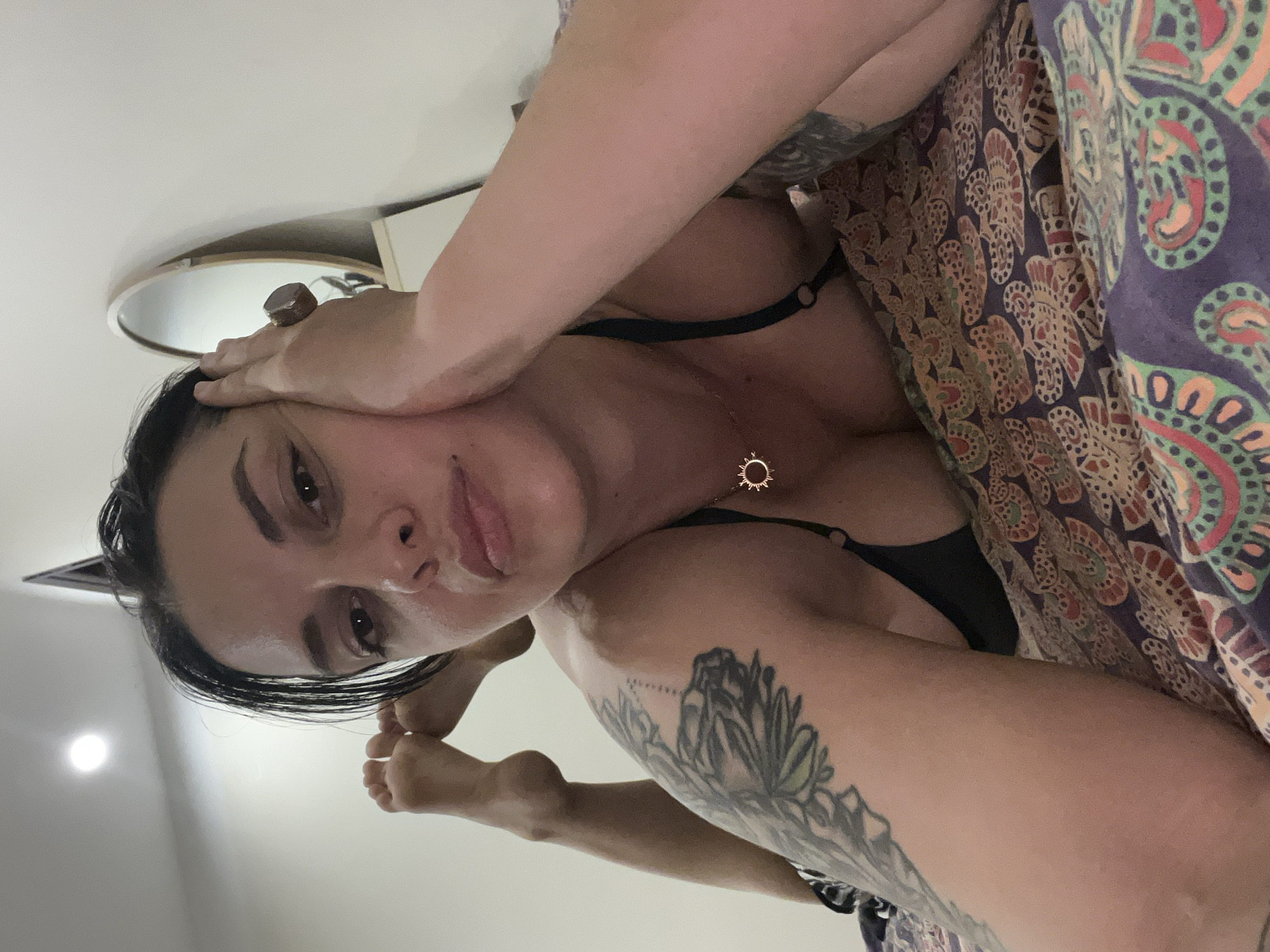 A close-up, low-angle shot captures a person lying down, with their head tilted downwards. The person has dark hair and is wearing a black bra and a necklace with a sun-shaped pendant. Their arms are positioned in a way that suggests they are holding their head or resting it. Visible tattoos adorn their arms. A patterned fabric, likely a blanket or cover, is draped over their body. The background appears to be an indoor setting with a ceiling light visible and a circular mirror reflecting some of the scene. The lighting suggests it could be daytime or artificial indoor lighting. The image is oriented sideways.