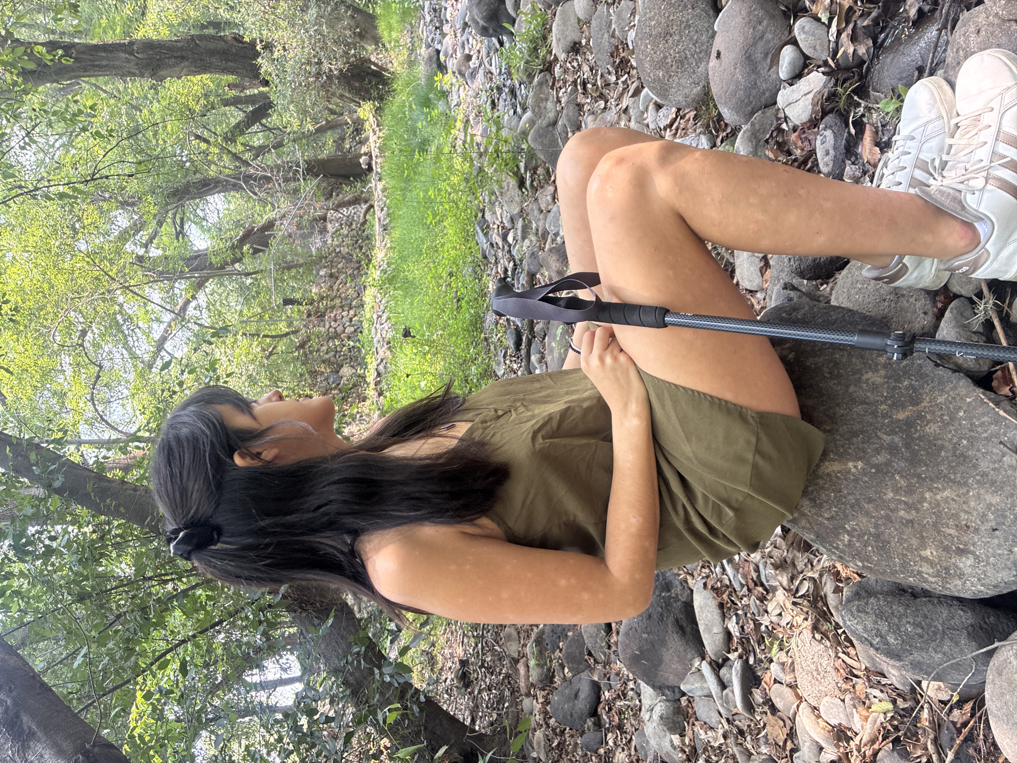 A woman is sitting on a large rock in a natural setting. She is wearing an olive green dress and white athletic shoes. She has long dark hair pulled back in a ponytail. She is holding a black trekking pole. The ground is covered with rocks, leaves, and vegetation. The location appears to be near a river or stream, surrounded by trees. The photo was taken in Cañada de Madero, Mexico.