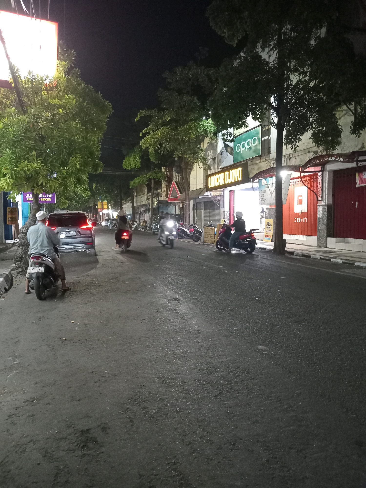 The image depicts a street scene in Gresik, Indonesia, at night. A dark asphalt road runs through the center, bordered by sidewalks and buildings. Several motorcycles are present, with two men on motorcycles visible in the foreground and middle ground. One man, wearing a white cap and grey shirt, sits on a motorcycle parked on the side of the road. Another man, wearing a helmet and a dark jacket, rides a red motorcycle further down the street. A dark colored SUV is parked on the left side of the road. Buildings with illuminated signs line the street, including "OPPO" and "LANGGAN DJOVO". Trees with green foliage are scattered along the sidewalk. The overall atmosphere is dimly lit, characteristic of a street at night.