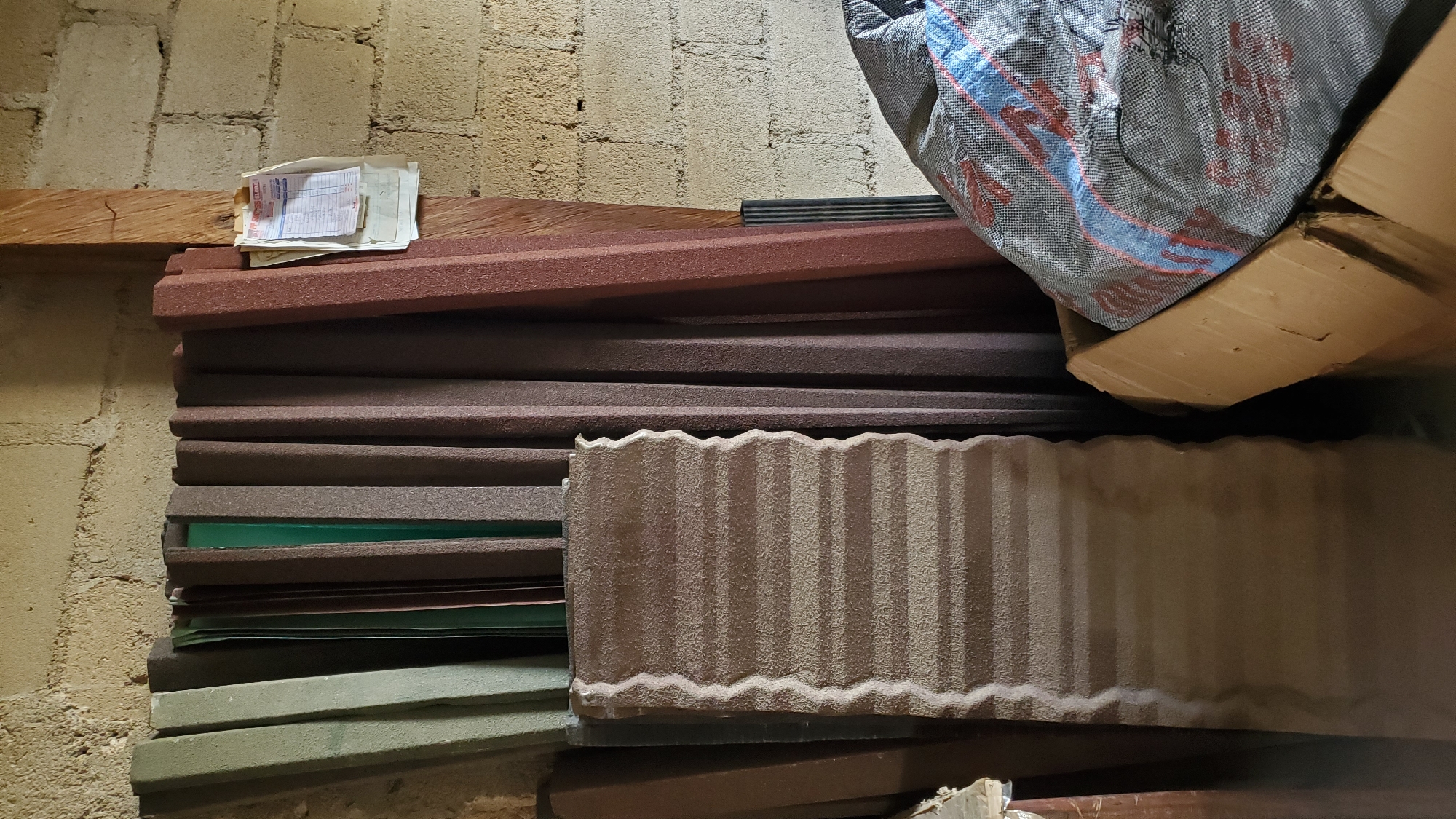 The image depicts a storage area, likely in Jalingo, Nigeria, with construction materials. In the foreground, a corrugated metal sheet with a textured, sandy finish is propped upright. Behind it, a stack of similar, but taller, metal sheets of varying colors, including green, brown, and reddish-brown, are leaning against a concrete block wall. To the right, a wooden post supports a small stack of papers, possibly receipts or invoices, attached with a nail. In the lower right corner, a large, grey woven sack with red and blue stripes is partially visible inside a cardboard box. The environment appears to be dimly lit and somewhat cluttered, suggesting a functional storage space rather than a public area.