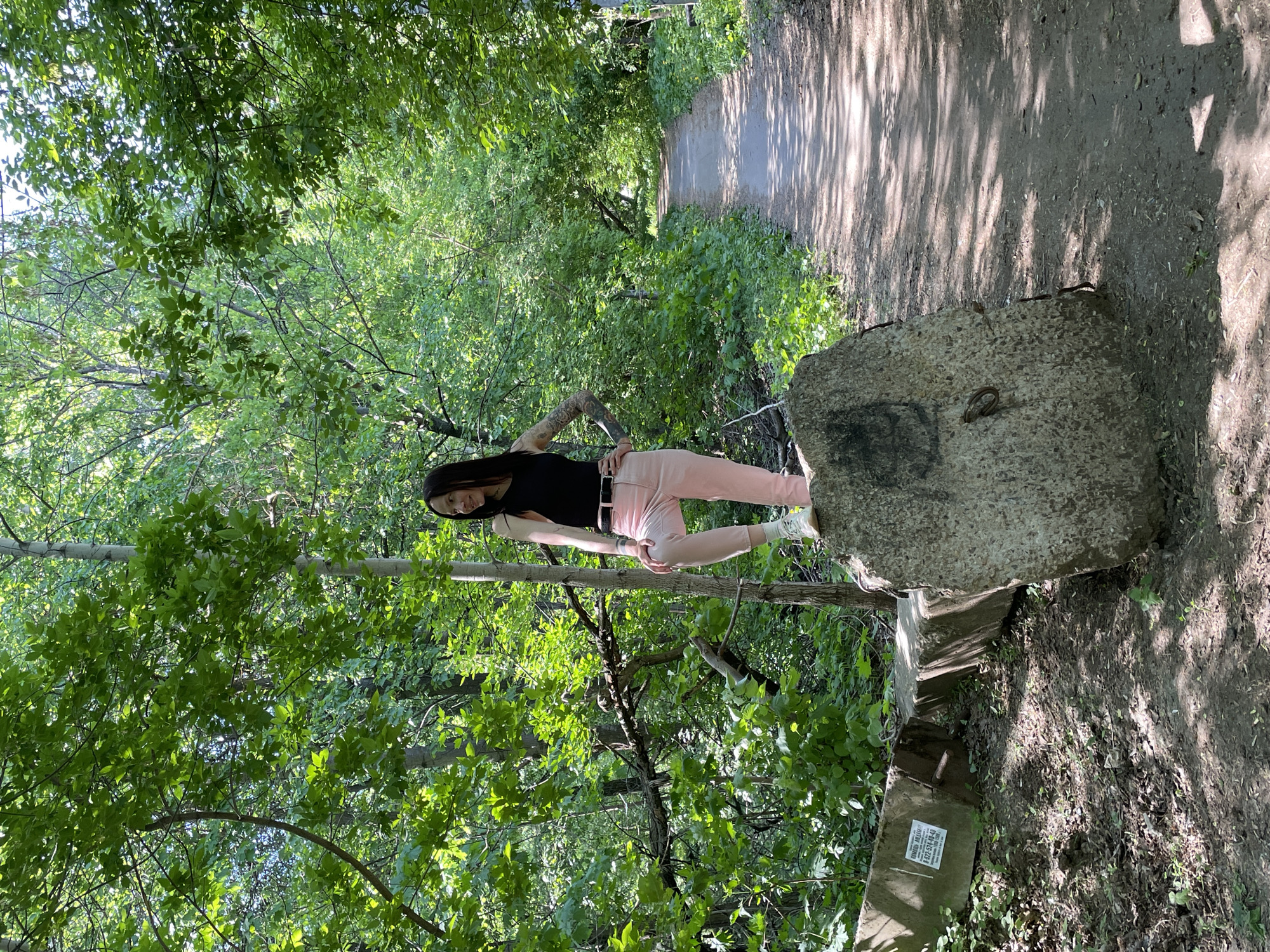 The image shows a woman standing outdoors in a wooded area in Oktyabr'skii, Russia. She is positioned between a thick, light gray concrete block with a metal ring affixed to one side, and a thin tree. The woman has long dark hair, is wearing a black top, pink pants, white sneakers, and a black belt. Her left arm is visible, displaying tattoos. She is standing on her right leg, her left leg bent at the knee and raised. The concrete block appears to be part of a larger structure or installation. The surrounding environment is filled with dense foliage and trees. A walking path is visible in the background.