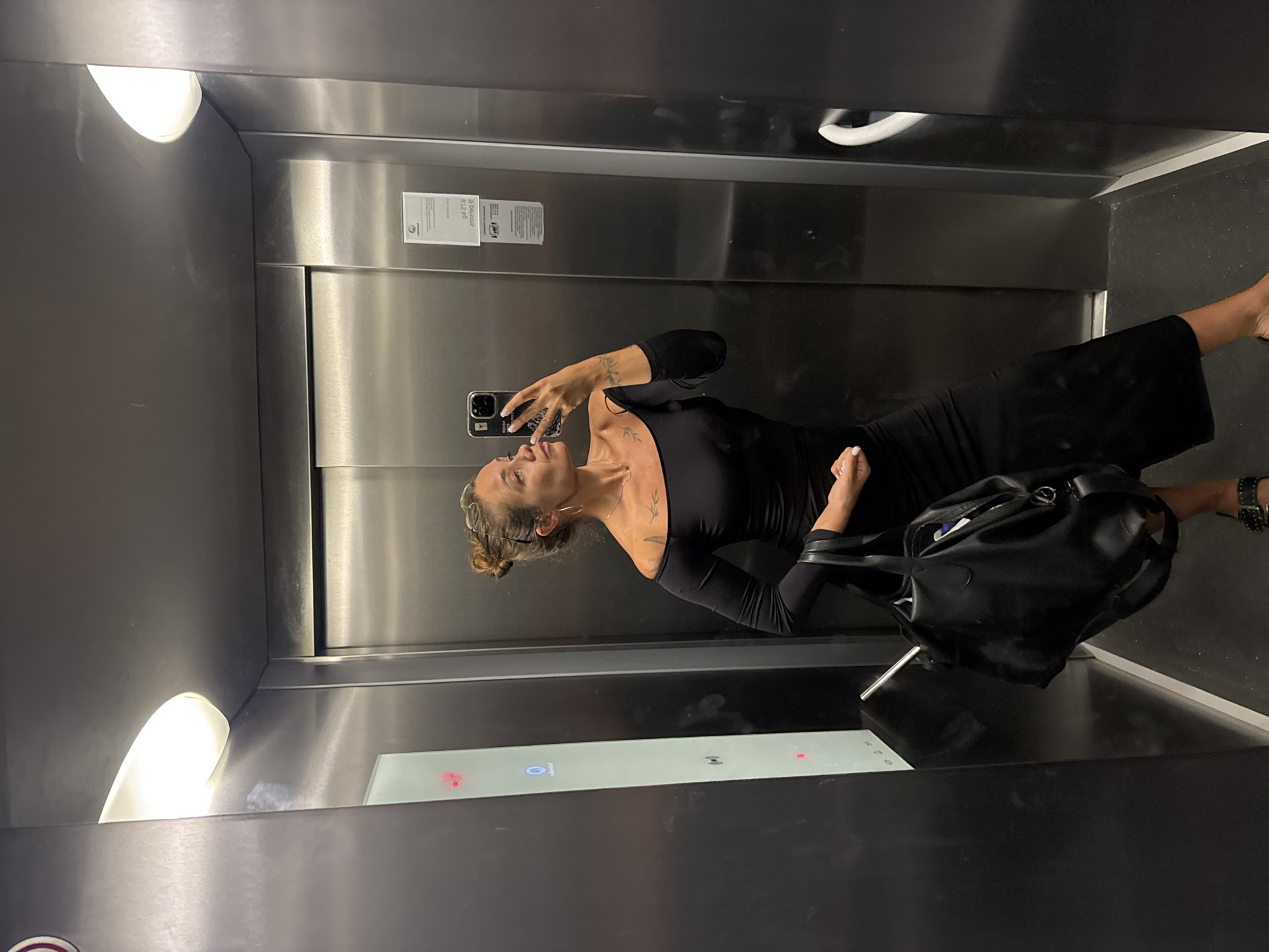 A woman stands in an elevator, taking a mirror selfie. She is wearing a black off-the-shoulder dress and holding a black handbag. Her hair is pulled up in a bun, and she has tattoos on her arm and shoulder. The elevator walls are made of brushed metal. There are two illuminated panels on the left wall, one with red buttons and another with what appears to be an intercom system. A small white information sign is visible on the right wall. The lighting in the elevator is provided by two overhead lights. The overall impression is that of a modern and functional elevator interior. The location is specified as Budva, Montenegro.