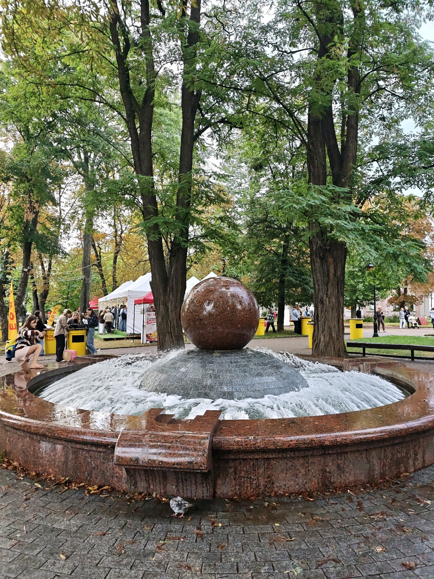 The image depicts a public fountain with a large, textured sphere at its center, surrounded by flowing water. The fountain's basin is constructed from reddish-brown granite. The scene is set in an urban park with mature trees exhibiting autumn foliage. In the background, several white tents suggest an outdoor event or market. People are visible in the midground, some near the tents and others in the foreground. A young woman in a striped sweater and light-colored skirt is crouching near the fountain. A pigeon is pecking at the wet paving stones in the lower right quadrant. The paving stones are laid in a cobblestone pattern. The overall impression is a lively, outdoor gathering in a park setting. This scene is located in the city of Vinnytsia, Ukraine.