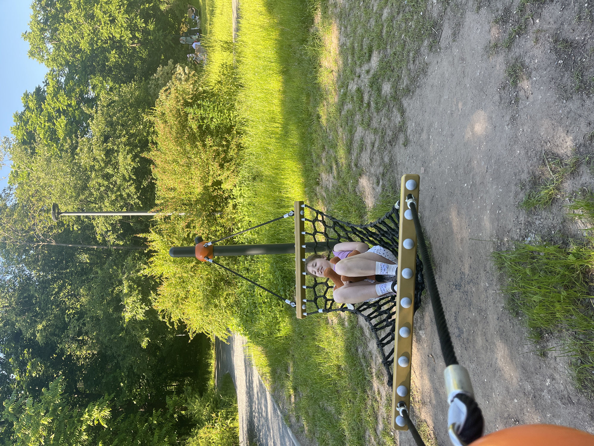 The image depicts an outdoor recreational scene within a park environment, in the city of Unknown, Unknown, under bright natural sunlight. The primary subject is a young pre-teen or early teenage girl with light brown hair, seated in a black mesh hammock-style swing suspended from a dark grey central pole with an orange top accent. She is wearing a purple tank top, white patterned shorts, and white socks, holding a reddish-brown stuffed animal, possibly a fox or small bear, in her lap. Her facial expression shows a relaxed smile with eyes slightly closed.
The swing features a yellow wooden bar at the bottom adorned with multiple white spherical elements, and is secured by black ropes. The ground immediately surrounding the swing consists of sparse dirt mixed with patches of green grass.
To the left of the swing, a paved pathway curves into the background, bordered by dense green bushes and trees. To the right, a vibrant green grassy area extends towards the background, where a group of approximately 4-6 individuals are visible relaxing on benches or blankets beneath tall trees. One person is distinctly visible in green clothing, and another in white. The background is dominated by a dense canopy of lush green trees and varied shrubbery, suggesting a well-vegetated park. The lighting indicates a clear day, likely late morning or afternoon, with distinct shadows visible on the ground and foliage. An orange spherical object is partially visible in the extreme lower left foreground, possibly another component of the playground equipment.