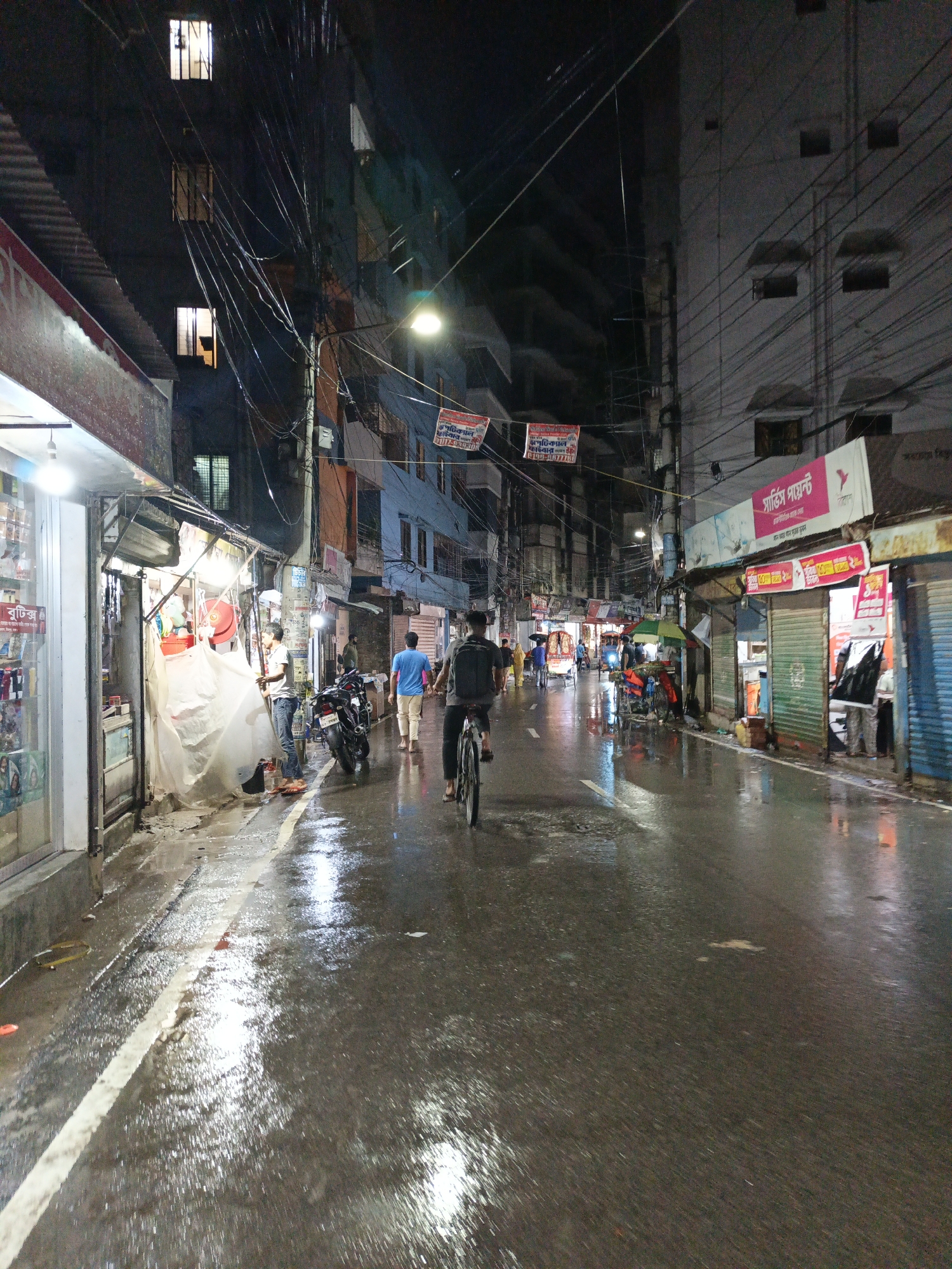 The image depicts a street scene in Dhaka, Bangladesh at night after a rain. The street is wet, reflecting the overhead streetlights. On both sides of the street are buildings with shops. Power lines crisscross overhead. In the foreground, a person is riding a bicycle. Further up the street, a man is walking, and several people are standing in front of a shop on the left. Another shop is open on the right side of the street.