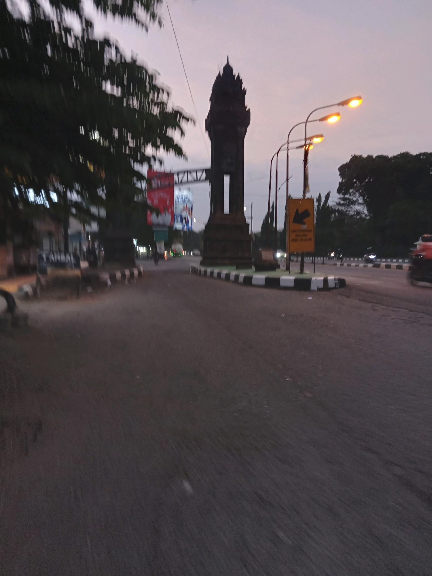 The image depicts a street scene in Bokoharjo, Indonesia, at dusk. A prominent dark, ornate tower stands at a roundabout, flanked by black and white striped curbs. Streetlights with yellow illumination line the street, casting a warm glow. In the background, trees and buildings are visible, some with lights on. A billboard with red and white advertising is also present. The road surface is asphalt, and there are signs of traffic with a blurred motorcycle rider visible. The overall atmosphere is subdued, indicating the transition from day to night.