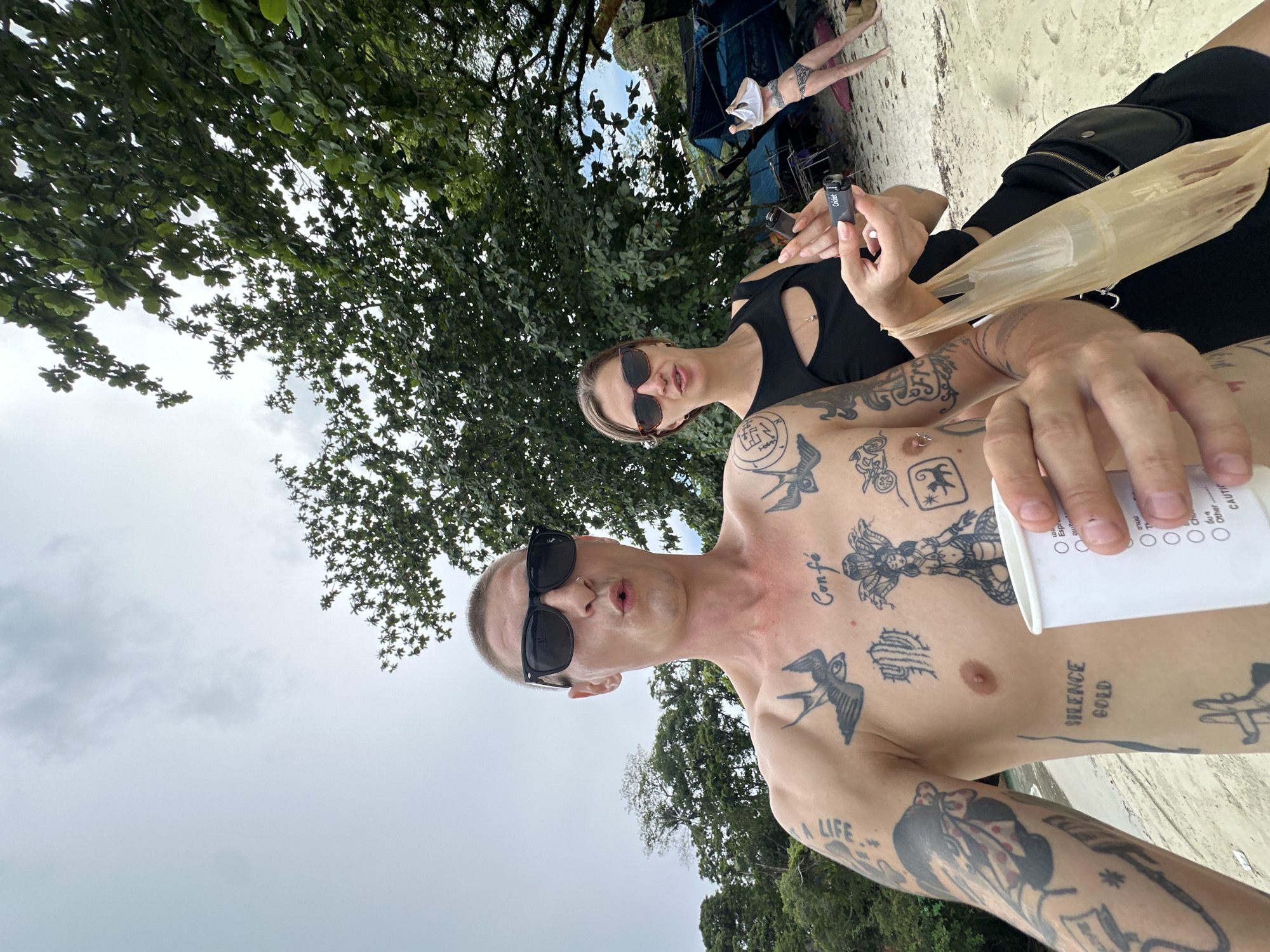 The image depicts a selfie taken outdoors on a beach, likely in Karon, Thailand, given the context. In the foreground, a man with numerous tattoos on his chest and arms is taking the picture. He is wearing black sunglasses and appears to be making a surprised expression. He is holding a white paper cup in his right hand. To his right, a woman is also visible. She is wearing a black top with a cut-out detail and sunglasses. She is holding two small cylindrical objects in her hands. Behind them, a beach environment is visible with sand, trees, and a cloudy sky. In the background, further down the beach, a woman in a bikini is standing, and there are some tents set up.