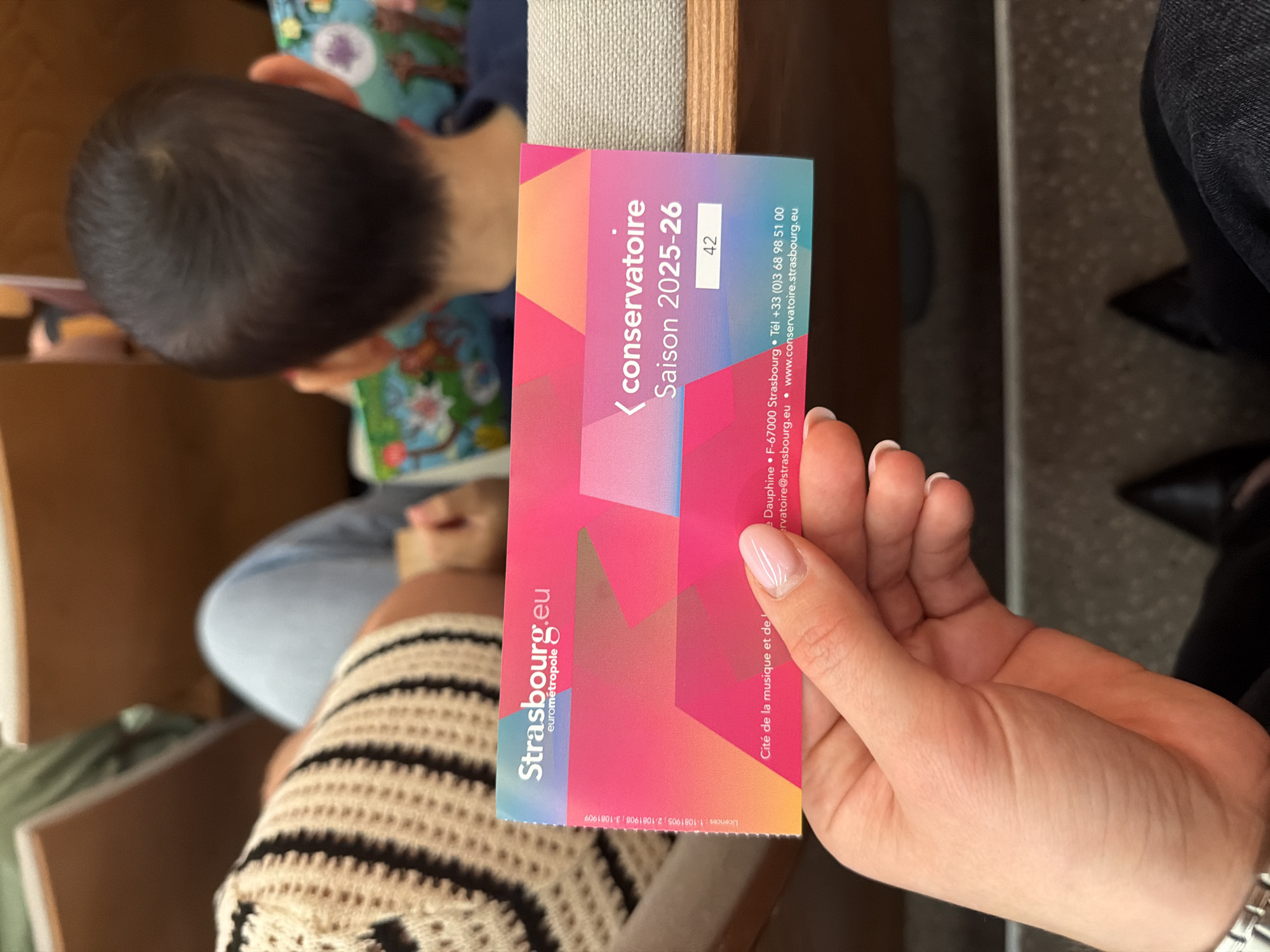 A close-up shot shows a person's hand holding a pink and blue ticket. The ticket reads "Strasbourg.eu eurometropole," and "conservatoire Saison 2025-26." Below this, the number "42" is displayed in a white box. At the bottom of the ticket, contact information is visible: "Cité de la musique et de l'opéra," "Dauphine, F-67000 Strasbourg," "Tél +33 (0)3 68 98 51 00," "conservatoire@strasbourg.eu," and "www.conservatoire.strasbourg.eu."
The ticket is held in front of a blurred background of people seated in what appears to be an auditorium or concert hall. To the left, a person wearing a cream-colored, knitted sweater with black stripes is visible. To the right, the back of a child's head, with short dark hair, is in focus. The seating appears to be upholstered in a light brown fabric, and wooden panels are visible.