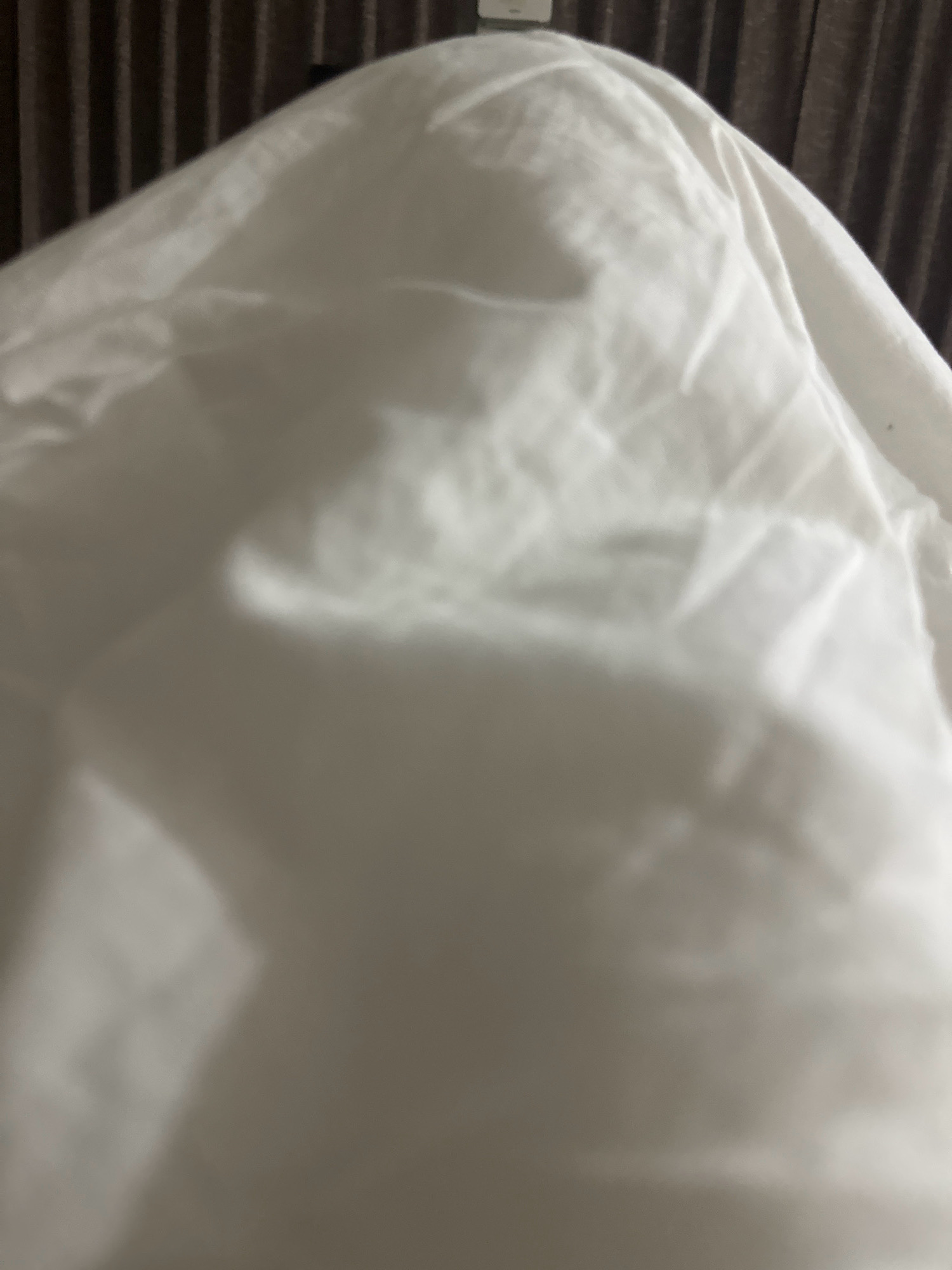 The image shows a close-up of a white fabric, possibly a bedsheet or curtain, draped over something unseen. The fabric has numerous folds and wrinkles, creating areas of light and shadow. In the background, vertical dark lines are visible, suggesting a textured surface such as blinds or a wall covering. The overall impression is of soft, diffused lighting. The location in Lalitpur, Nepal, cannot be determined from the visual information presented.