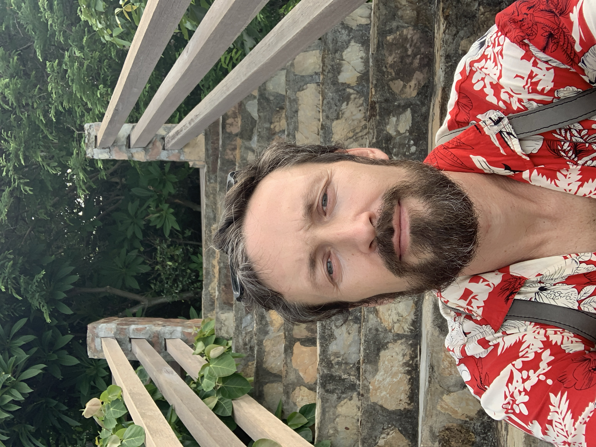 A medium shot photo shows a man with a beard and graying hair taking a selfie. He is wearing a red and white floral print shirt and sunglasses are perched on his head. He is sitting on a stone staircase, with wooden railings on either side. Lush green foliage and trees are visible in the background. The location is Baan Chaweng Noi, Thailand.