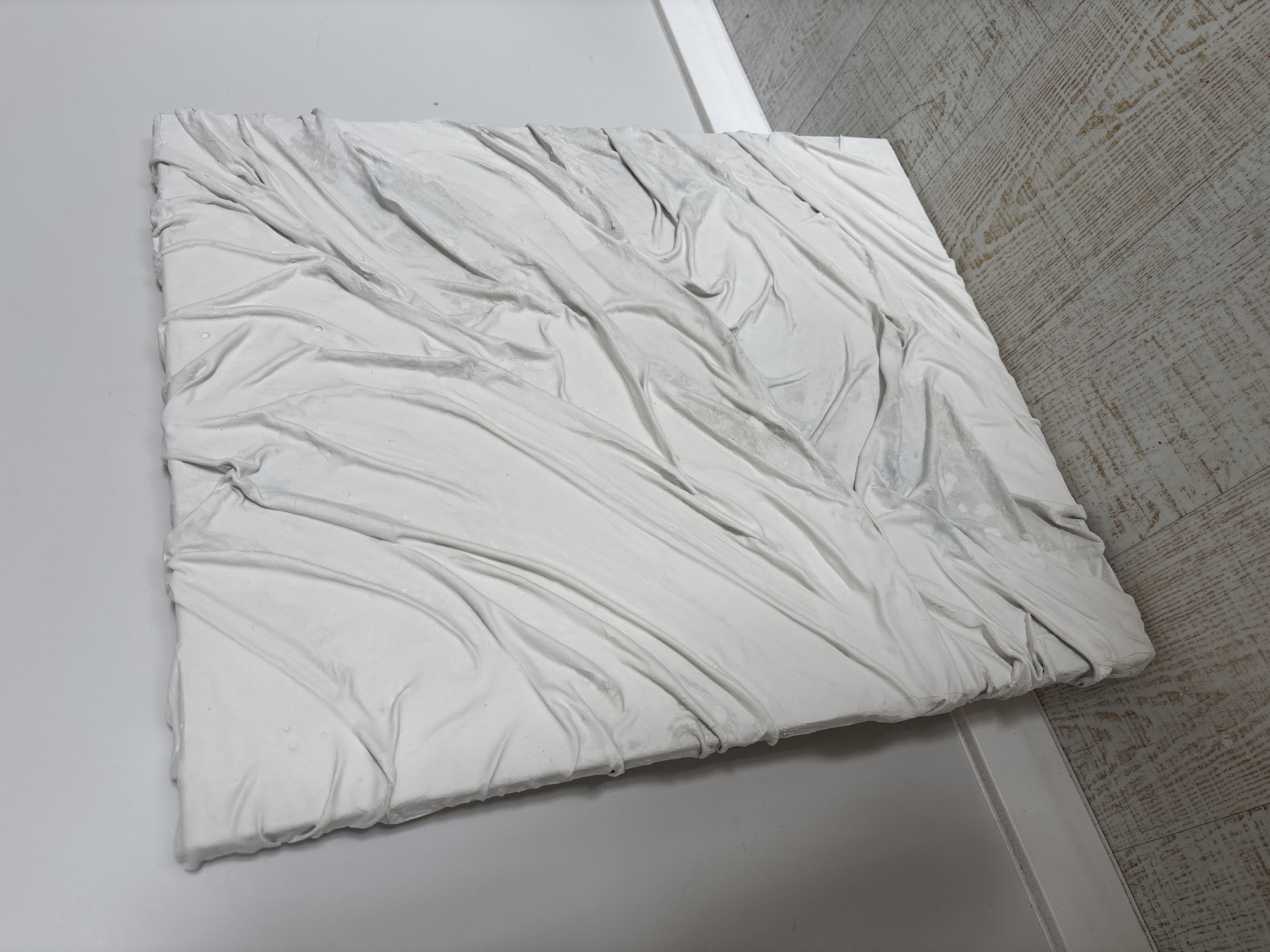 The primary subject is a square-shaped, three-dimensional artwork, likely a relief sculpture or a textured canvas, predominantly white. Its surface is covered in a material that is intricately creased, folded, and wrinkled, resembling draped or crumpled fabric that has been hardened or cast. The texture creates numerous peaks and valleys, casting subtle shadows that highlight its dimensionality. The overall appearance is abstract and monochromatic, with some slight variations in tone due to light and shadow, and possibly some minor discoloration or imperfections.
The artwork rests on a light-colored, flat surface, possibly a floor or a large table, which occupies the left and upper parts of the background. To the right and extending into the distance, there is a light-colored wooden floor with a distinct horizontal grain pattern. A white, narrow trim or baseboard delineates the boundary between the wooden floor and the other flat surface. The setting appears to be indoors, likely a studio, gallery, or a modern interior space, with even, diffused lighting that illuminates the textures of the artwork without harsh shadows. No people or activities are depicted, and there is no visible text. The image captures the artwork in a static, still-life presentation. The location context provided is Krasnoyarsk, Russia, though no specific visual cues in the image identify this city.