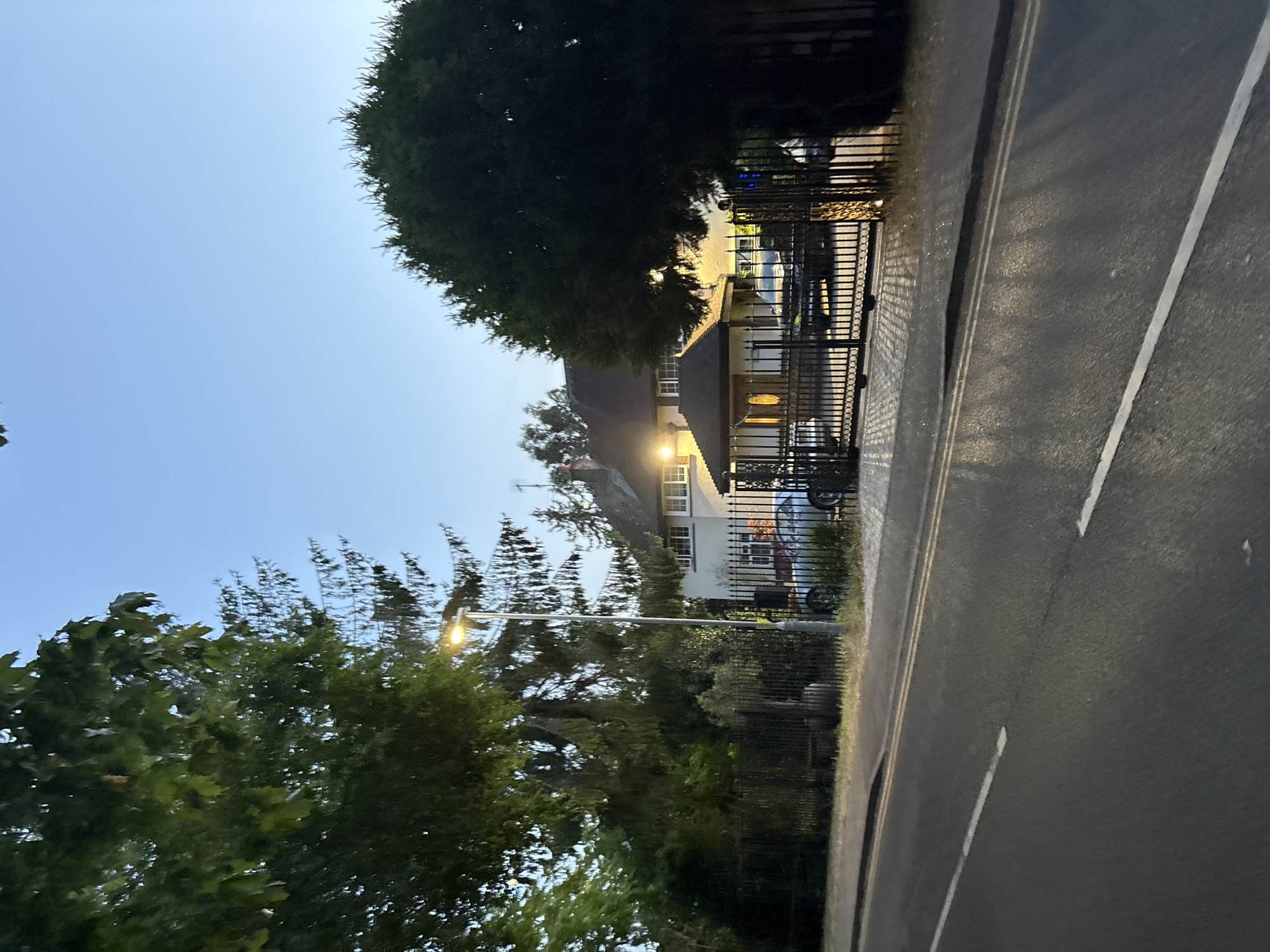A twilight view of a residential property at 348 Springfield Road, Chelmsford, United Kingdom. The scene is captured from across the road, which has white lines and appears to be asphalt. To the left, a large tree with green foliage dominates the foreground, with a street light and tall coniferous trees behind it. The property is behind a black metal fence. A house with a pale facade and several windows is visible, with a prominent pitched roof. In front of the house, there is a covered entrance with intricate metalwork and a dark roof. Two silver cars are parked within the property, visible through the fence and gate. A large dark evergreen tree is situated to the right of the property, obscuring some of the view. The sky is a clear, pale blue, indicating dusk or dawn.