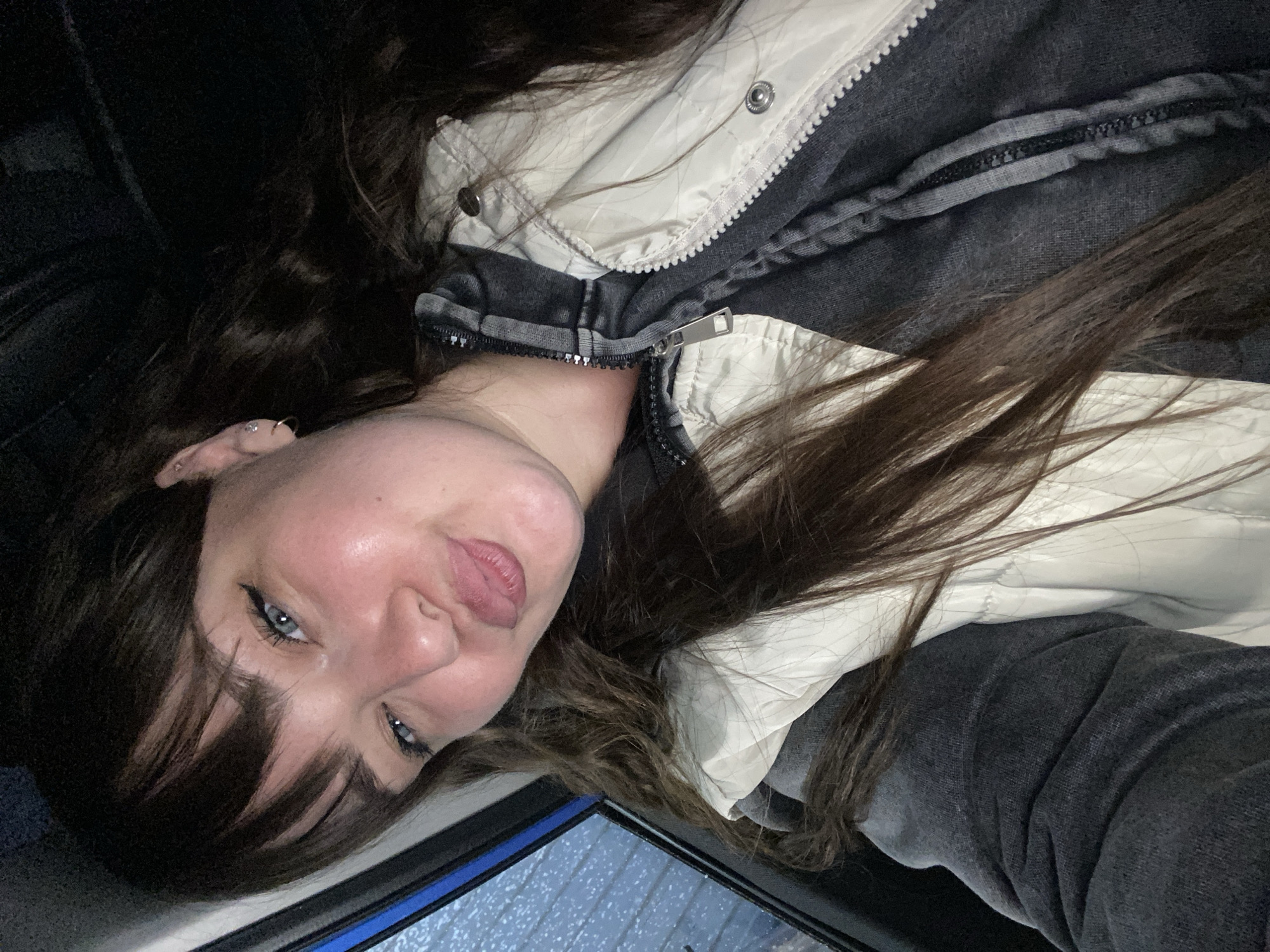 A selfie shows a fair-skinned woman with long brown hair, bangs, and blue eyes looking up and to the left, with a slightly puckered expression. She is wearing a jacket that has a light beige quilted outer layer and a dark gray inner layer, with a visible zipper. A window with rain droplets is visible in the lower right corner. There is no information given that implies this photo was taken in Votkinsk, Russia.